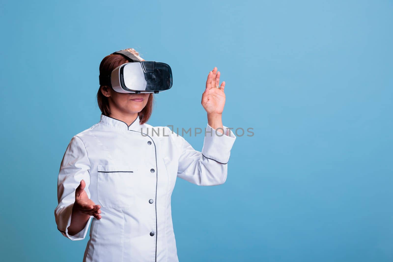 Cheerful cook woman using virtual reality headset while cooking healthy dinner in studio with blue background. Asian chef preparing culinary recipe for restaurant. Food industry concept