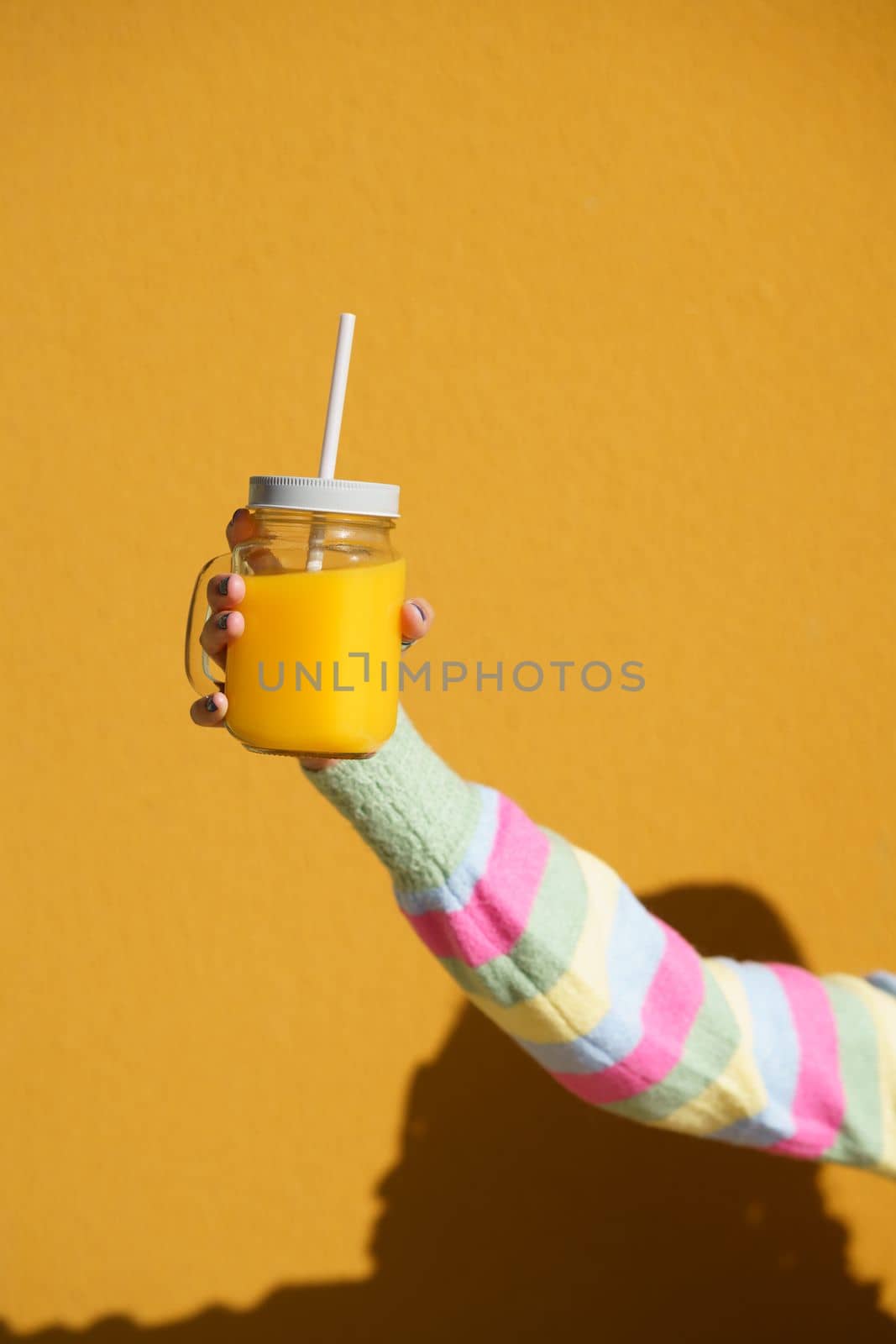 Female hands holding a glass tumbler with lid and straw, to take away, filled with fresh orange juice. Yellow background.