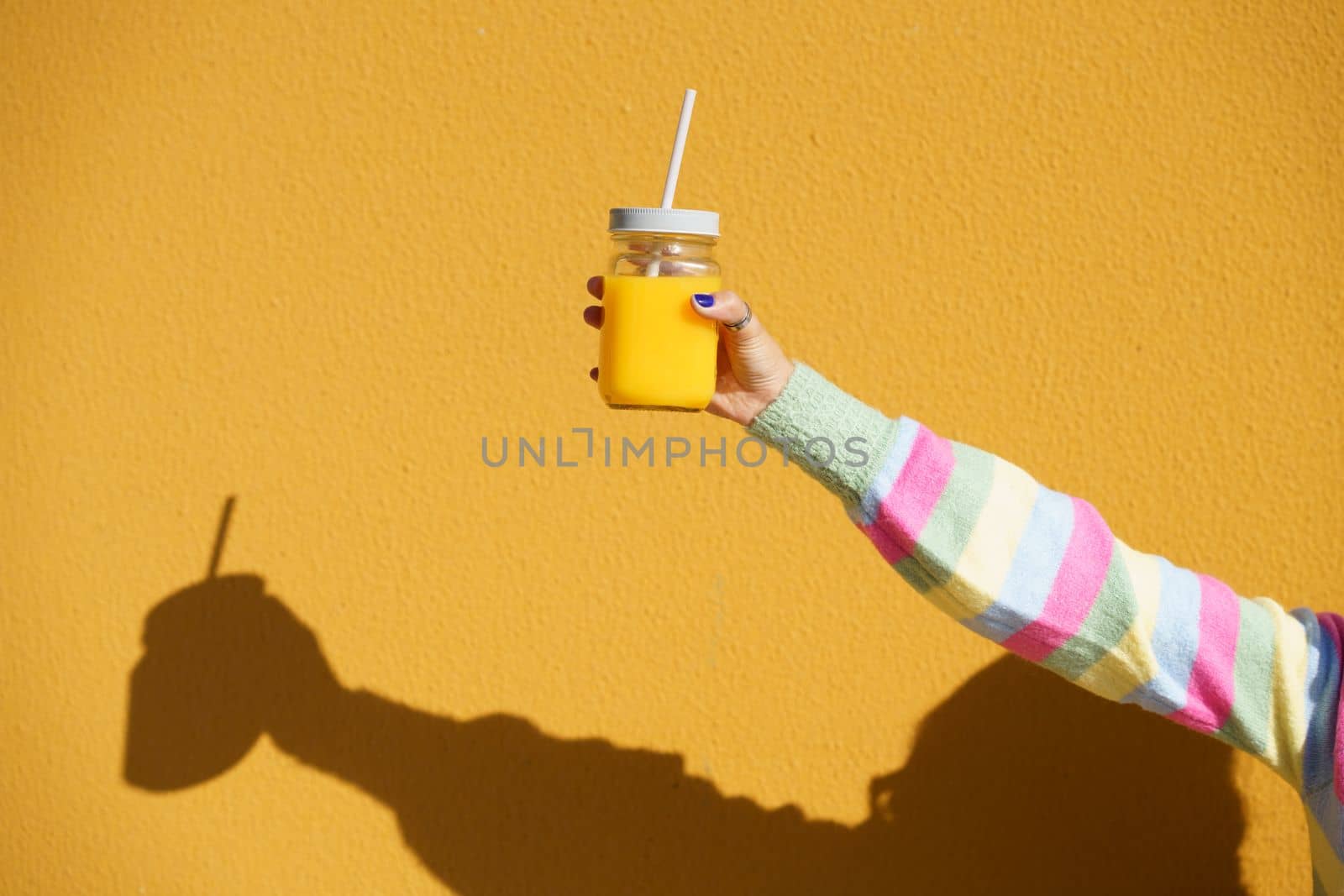 Female hands holding a glass tumbler with lid and straw, to take away, filled with fresh orange juice. Yellow background.