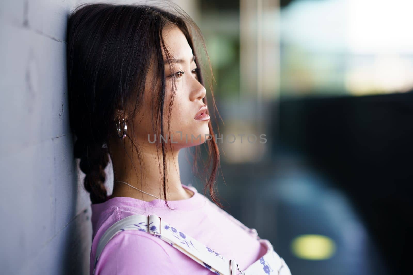Young Asian female in casual clothes with braid leaning on wall and looking away on city street in daytime
