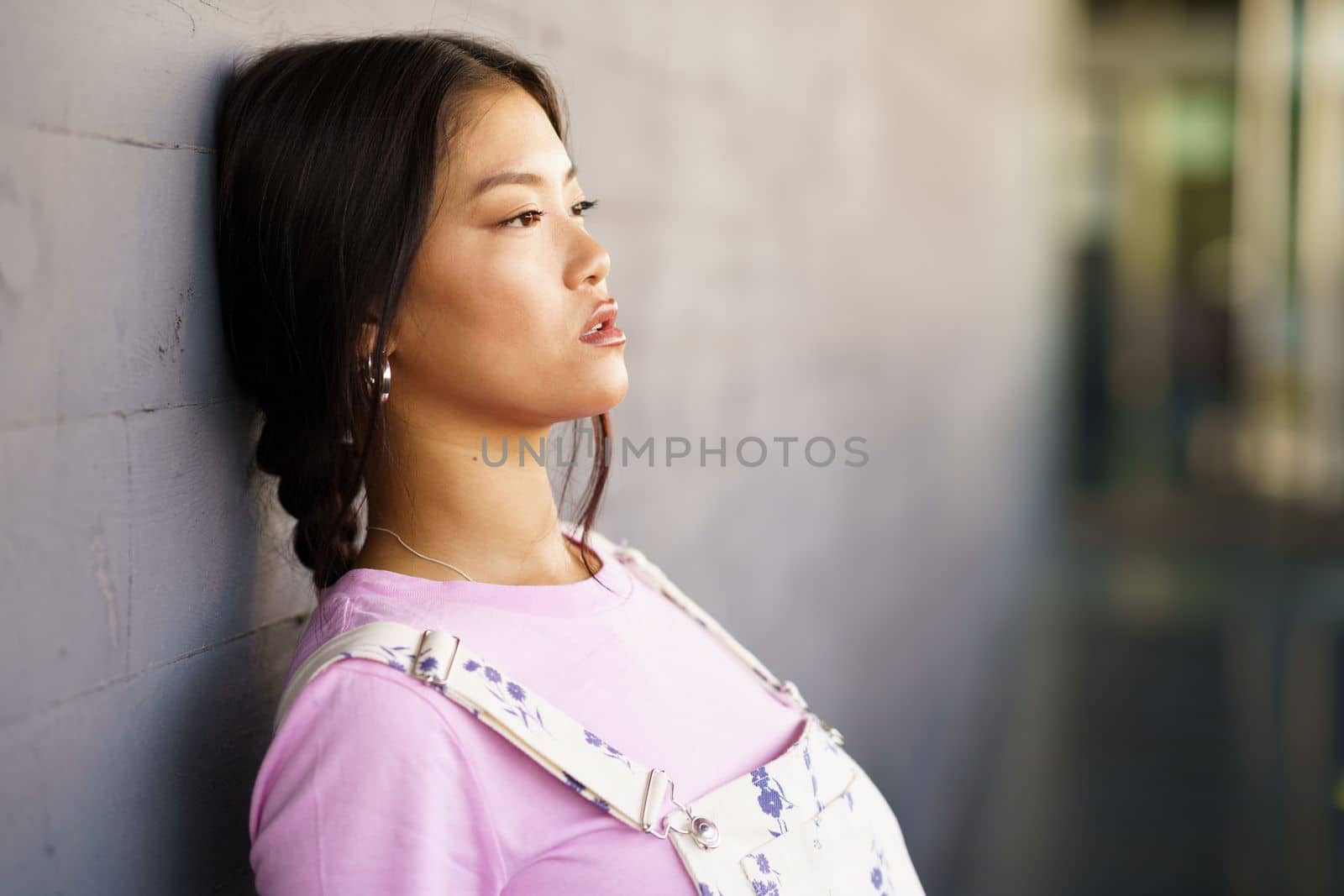 Young Chinese woman with a lost look and a serious expression leaning against the wall of a modern office building. Female wearing casual clothes.