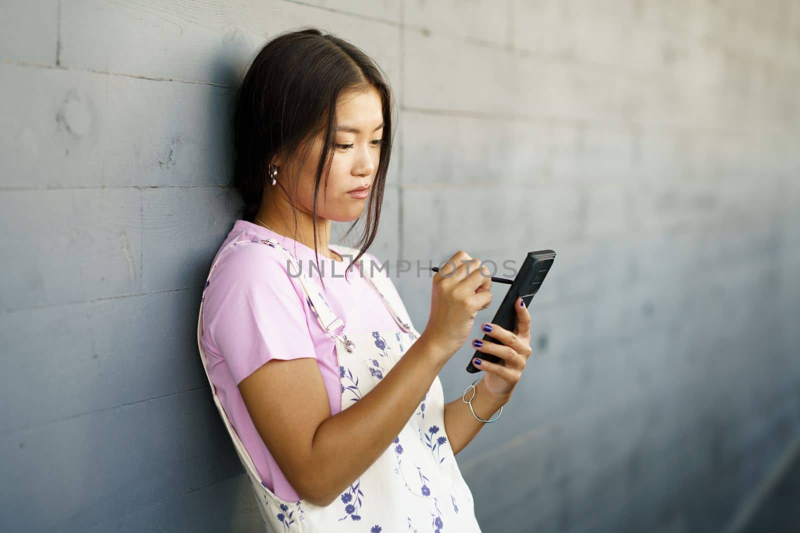 Serious Chinese girl using her smartphone with a pen or stylus,outdoors Asian female wearing casual clothes.