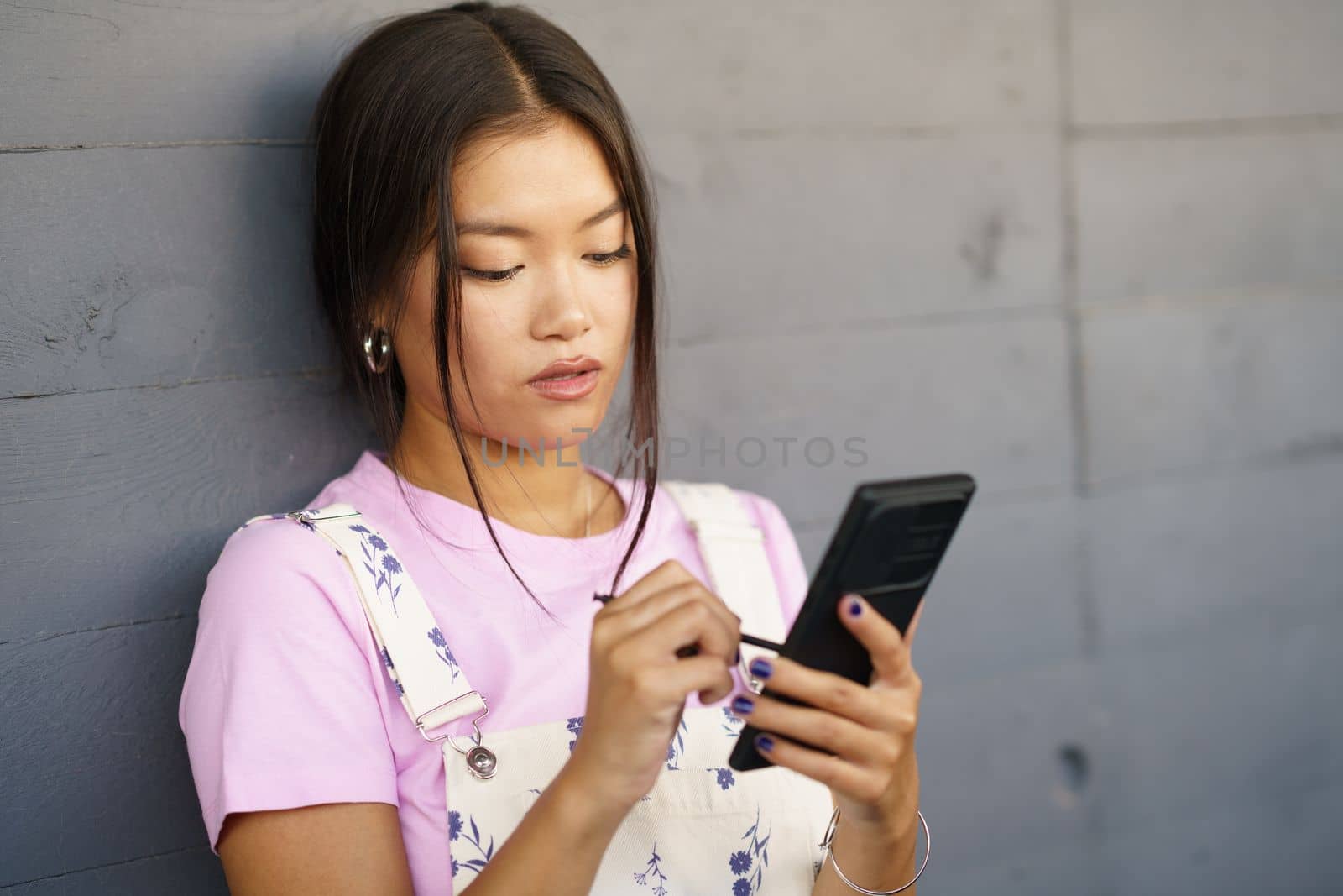 Serious Asian young woman using her smartphone with a pen or stylus,outdoors. by javiindy