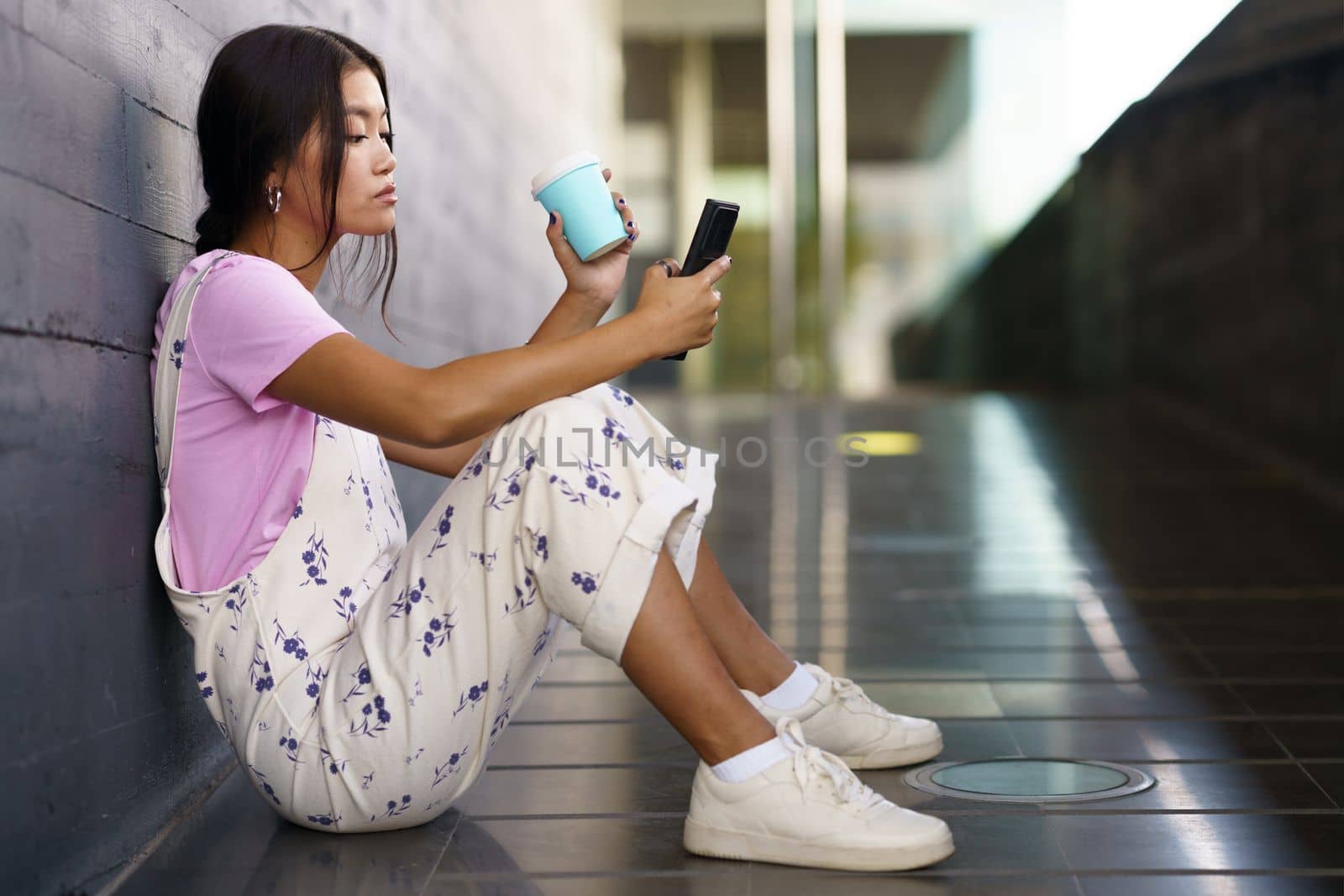Full body young Asian woman in stylish clothes with cup of coffee to go siting on slope outside modern building and browsing smartphone in daytime