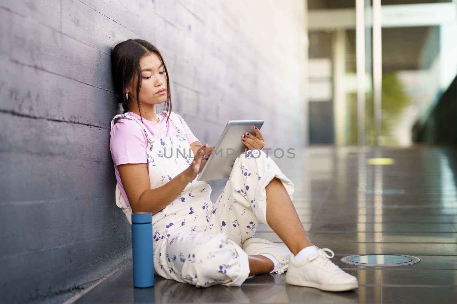 Asian girl consulting her digital tablet while taking a coffee break with a metal thermos.