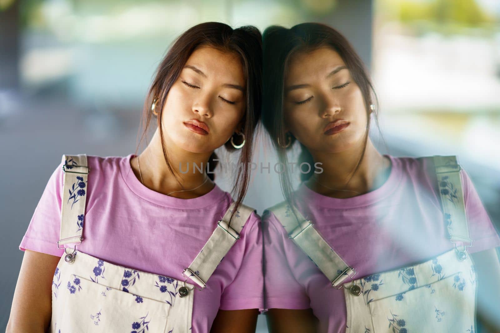 Calm stylish Asian female closing eyes and leaning on reflective glass wall on sunny day on city street