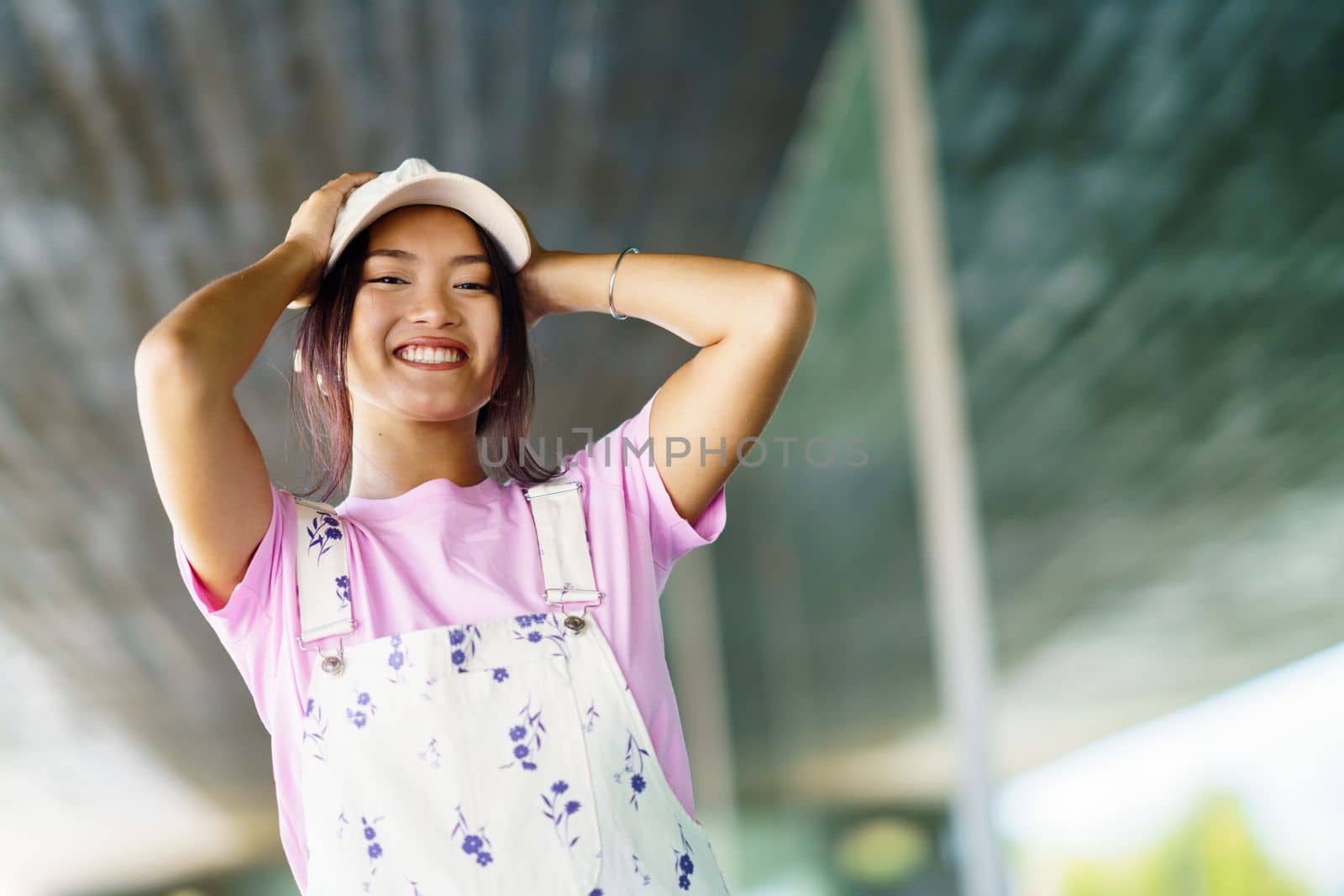 Low angle of positive young Asian female looking at camera smiling with happiness. Chinese woman in casual clothes and cap, near a modern building.