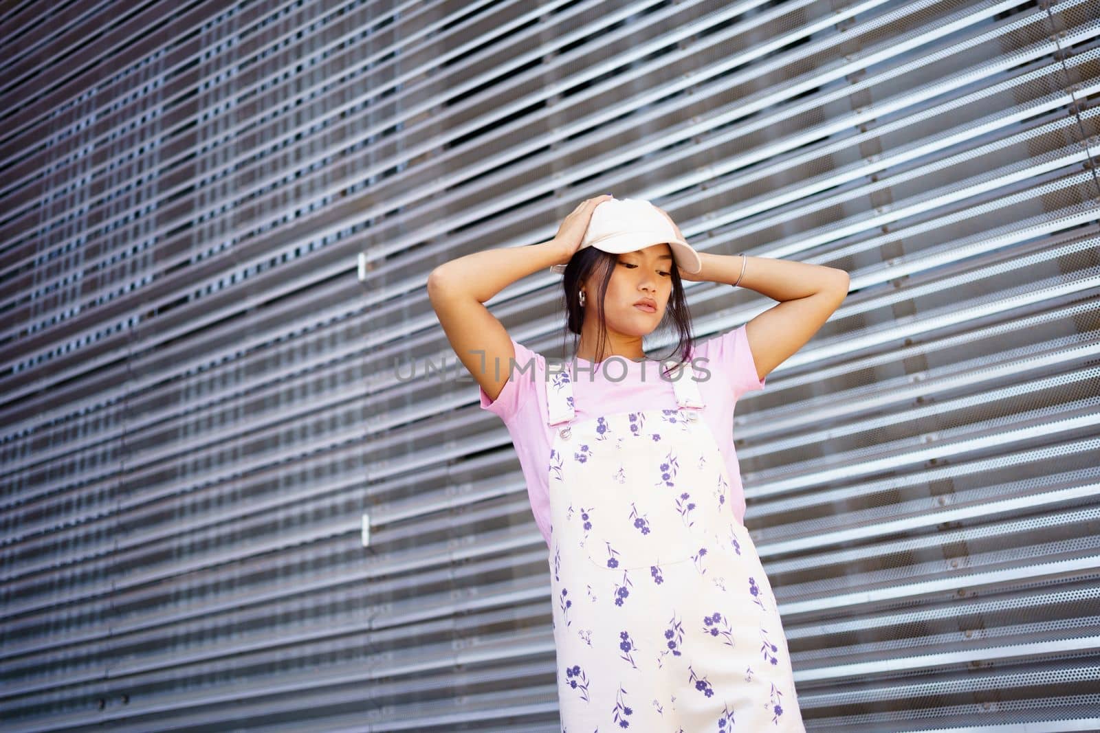 From below young Asian female in stylish clothes and cap touching head and looking down while standing near metal wall of modern building