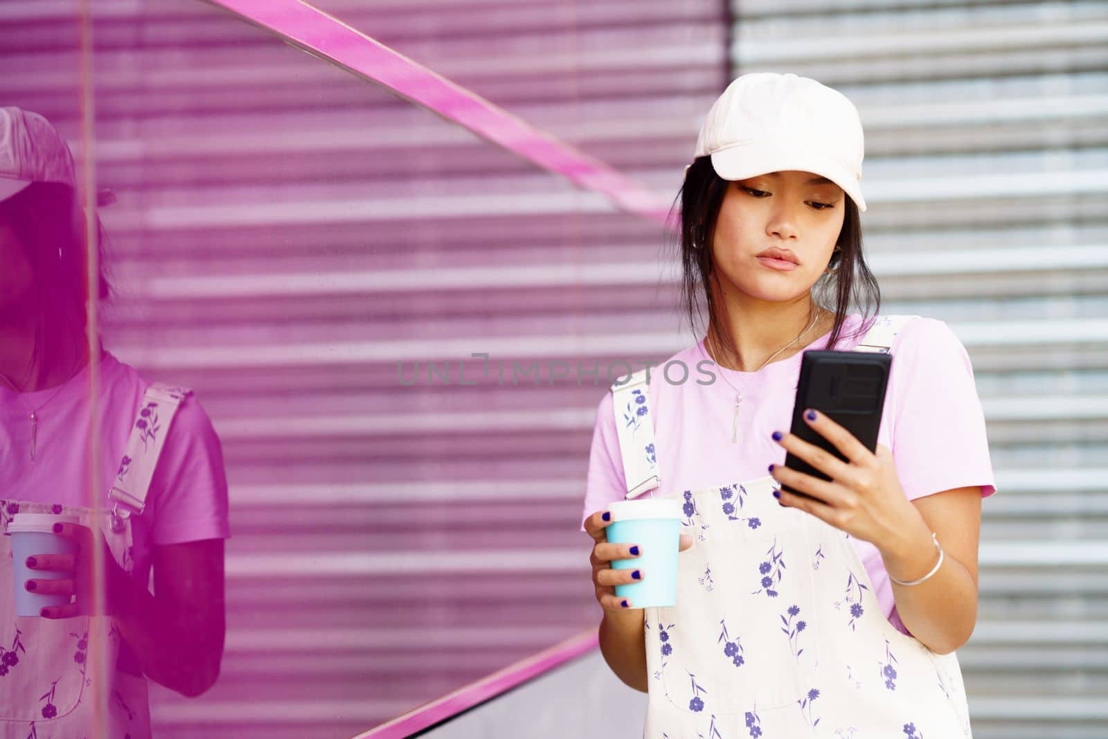 Young Asian female in stylish clothes and cap with takeaway coffee, browsing social media on cellphone while standing near pink glass wall of modern building on street