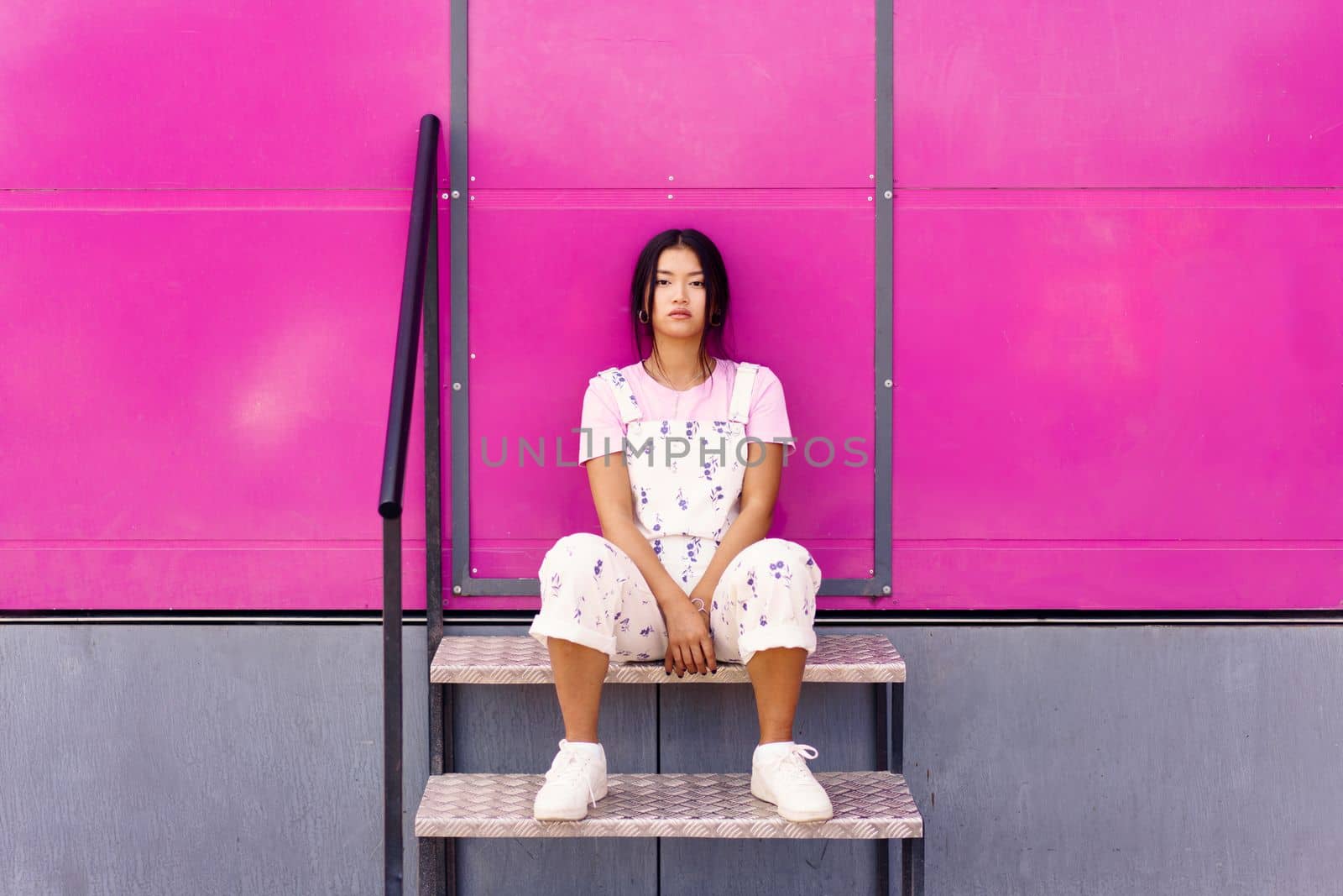 Full body stylish Asian female with dark hair sitting on metal stairs against pink wall and looking at camera on street