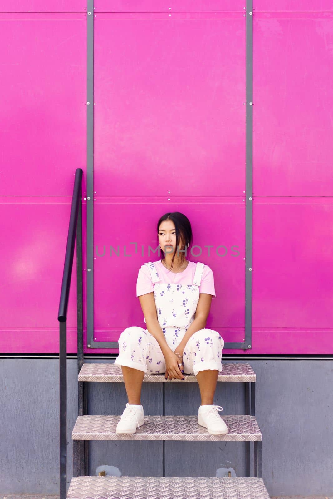 Full body tranquil young Asian female in stylish clothes sitting on metal steps against pink wall and looking away