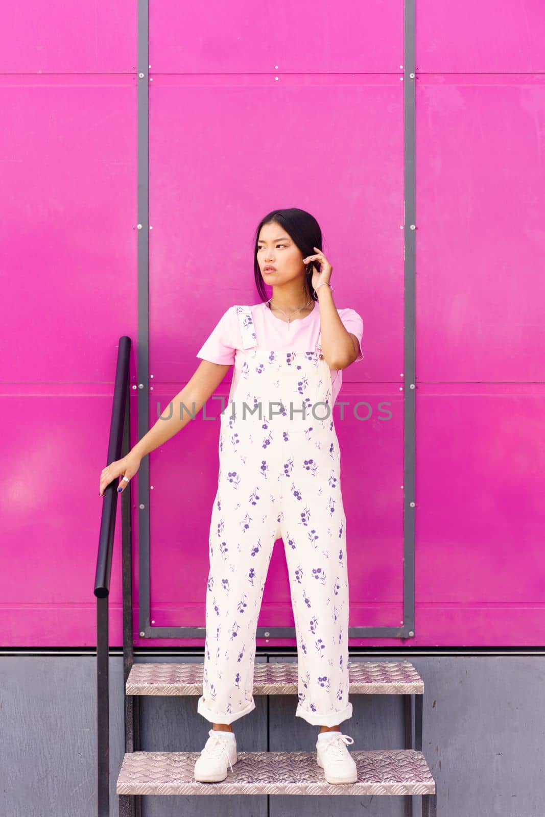 Chinese woman with blank stare and serious expression standing against pink wall of modern building. by javiindy