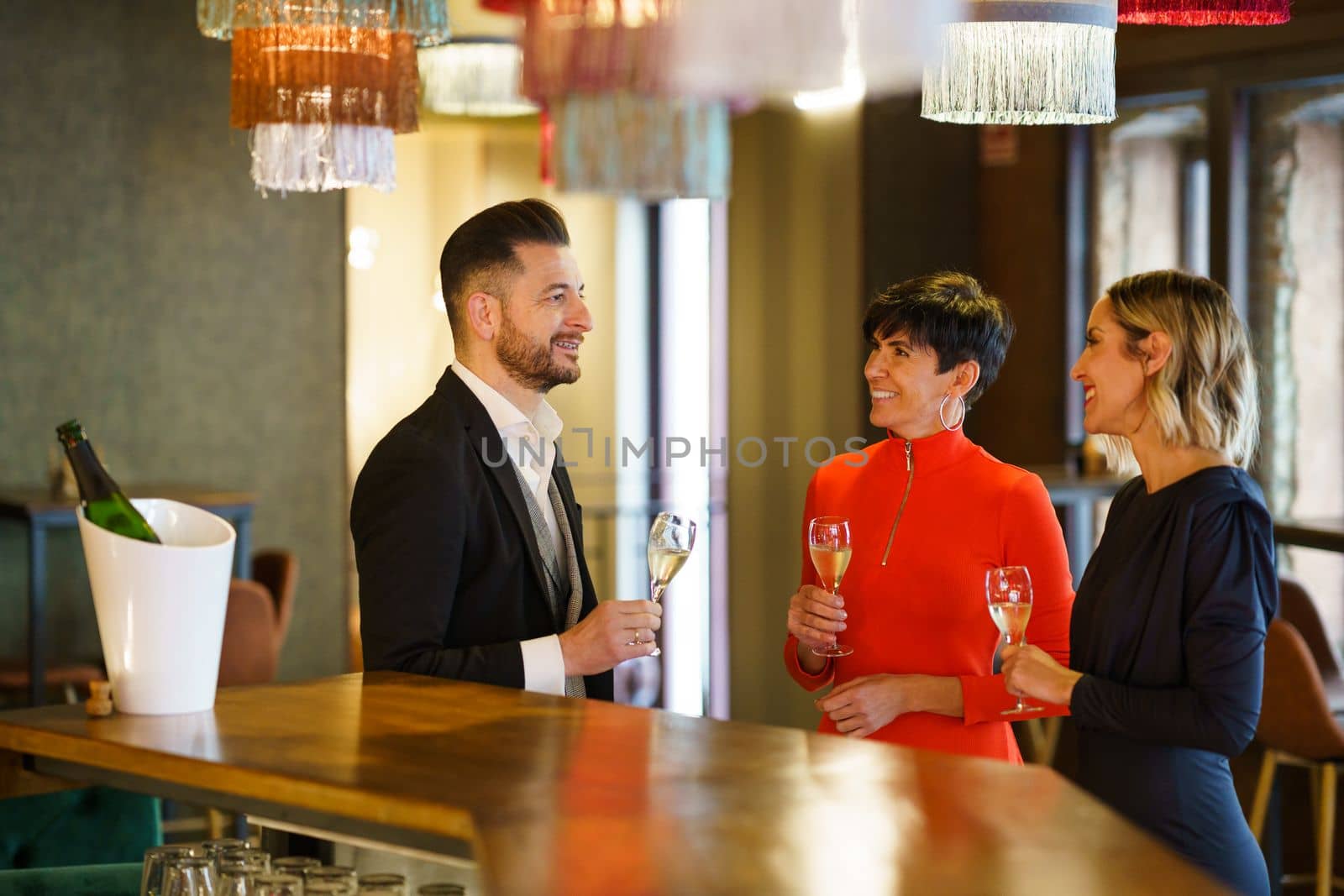 Group of elegant friendly people gathering at counter with glasses of champagne while enjoying meeting in bar