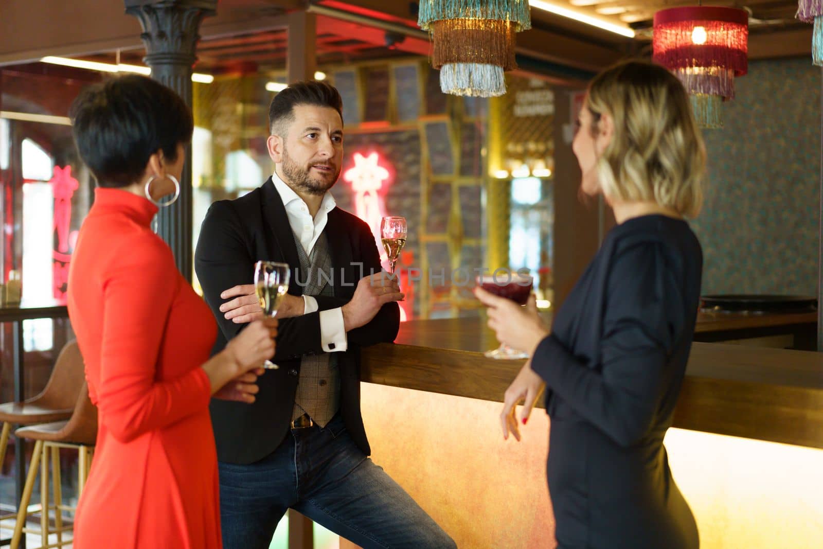 Group of elegant friendly people gathering at counter with glasses of champagne while enjoying meeting in bar