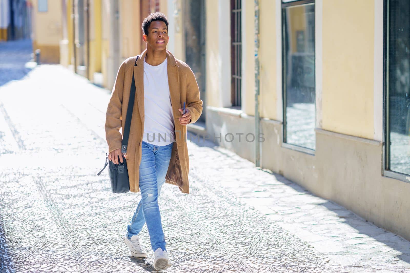 Young black man walking down the street carrying a briefcase and a smartphone. Cuban guy in urban background.