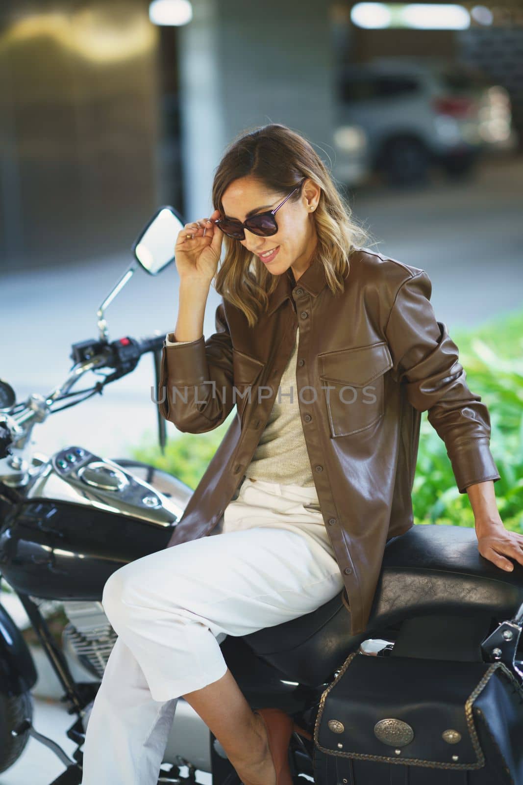 Stylish woman in sunglasses touching hair and looking down while sitting on motorbike on sunny day on street