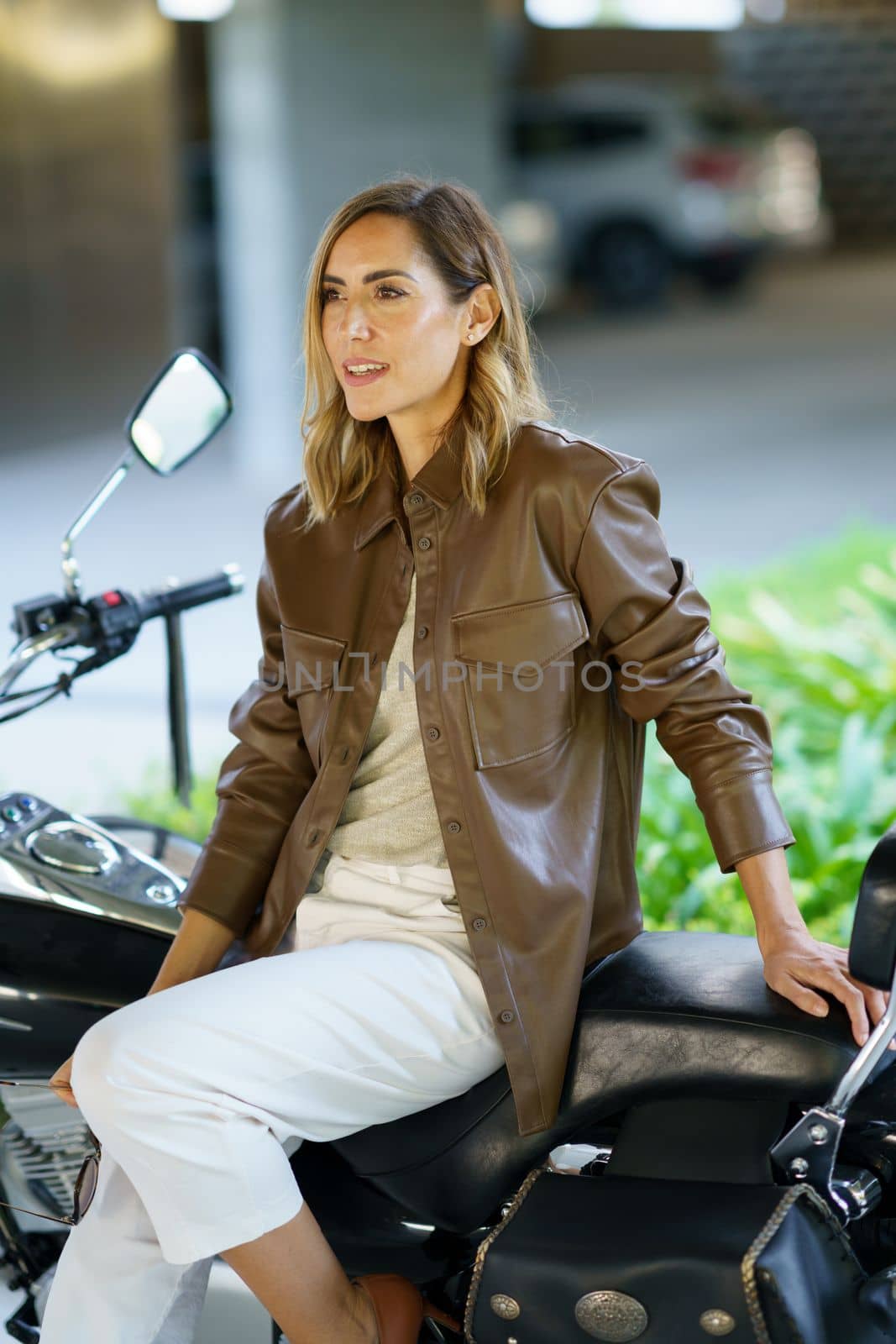 Adult female biker in stylish clothes looking away while sitting on motorcycle on blurred background of street