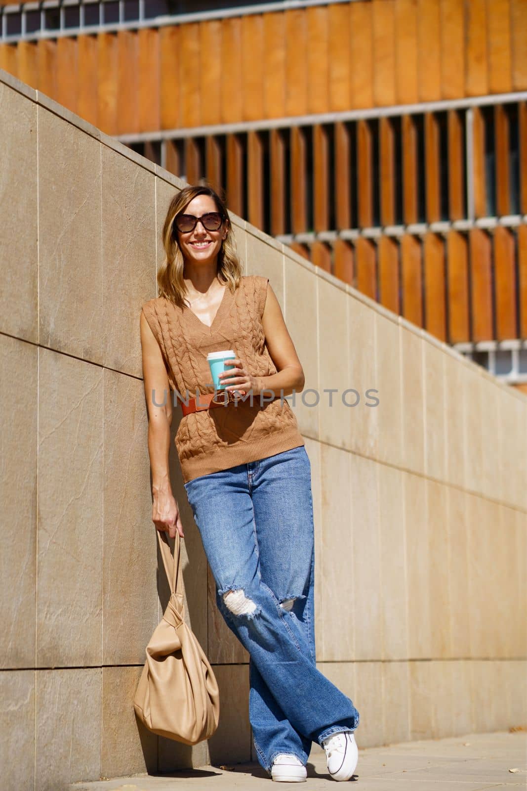 Full body adult woman in stylish clothes with bag and coffee to go leaning on wall and looking away on city street