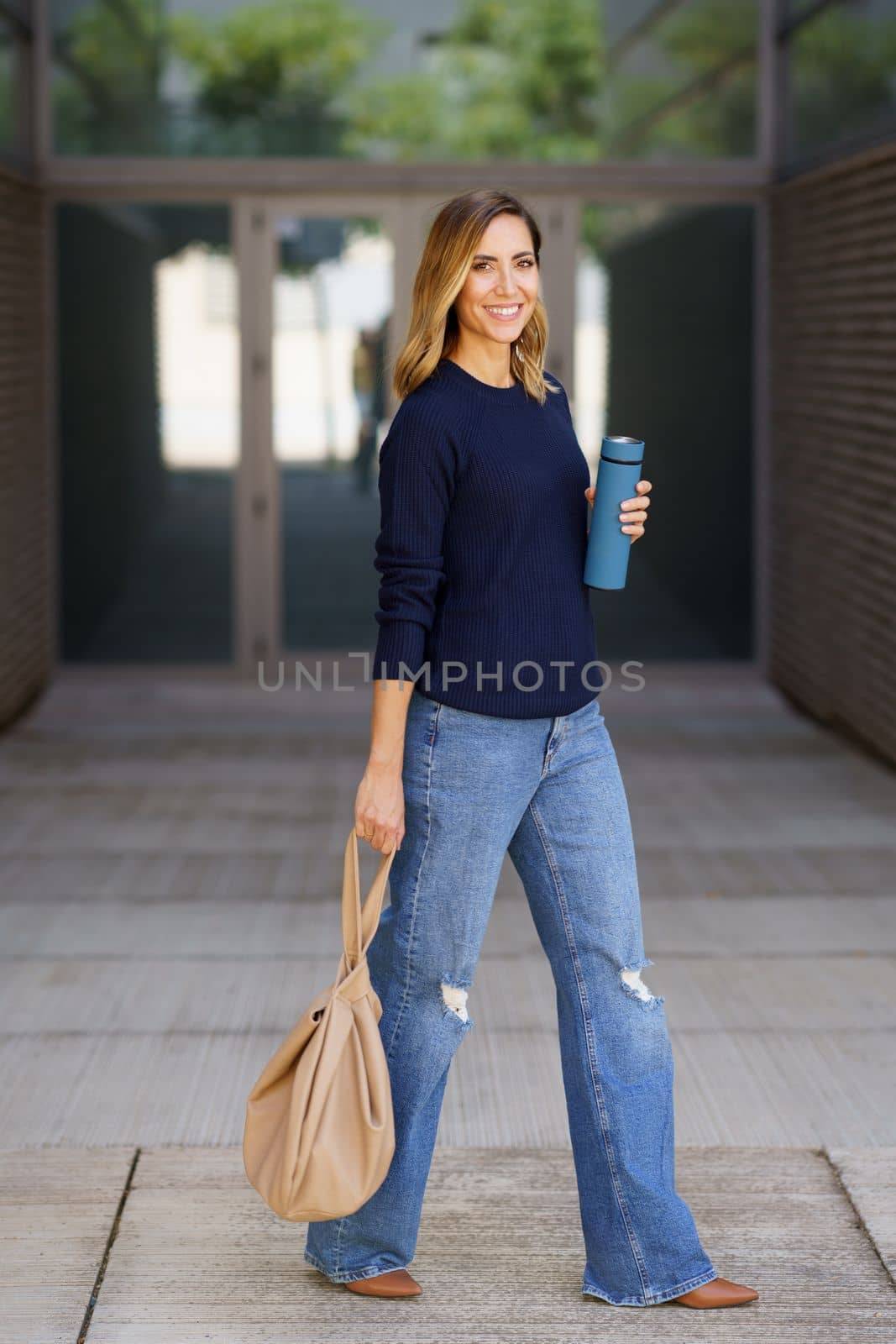Full body positive woman in stylish jeans and sweater with thermos and bag strolling on pavement near contemporary building and looking at camera with smile