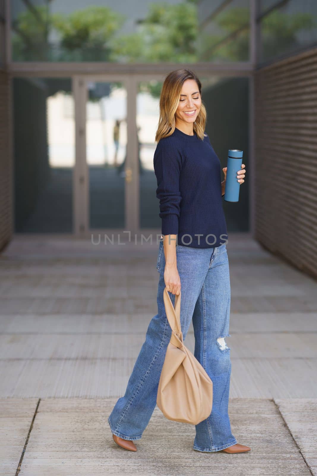Full body positive stylish woman with thermos and bag closing eyes and smiling while walking on pavement outside modern building