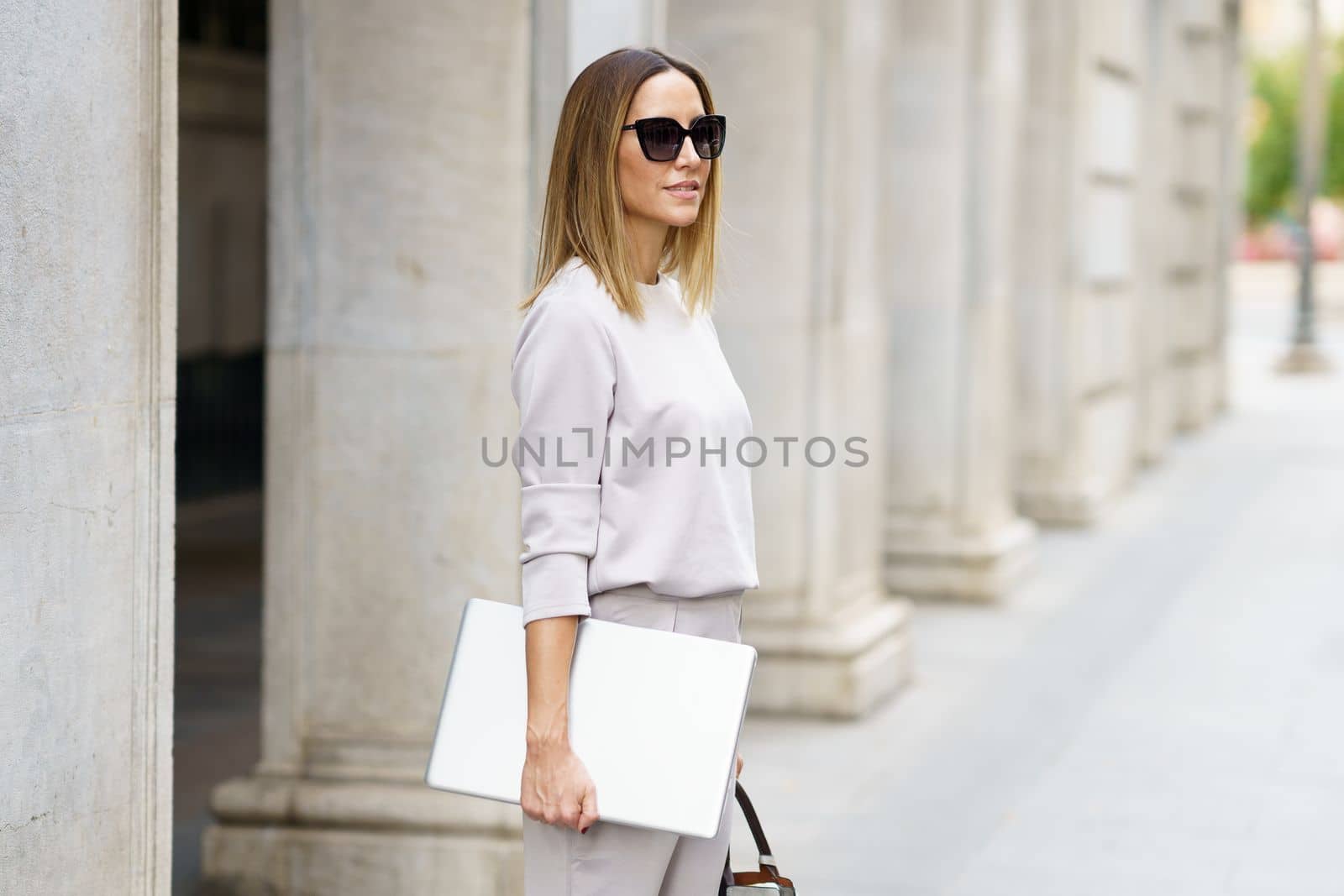 Side view of positive woman in gray outfit and sunglasses standing on paved street near building with columns and holding laptop while looking away