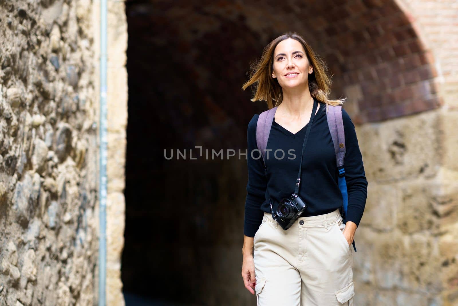Delighted young female tourist in casual clothes with backpack and photo camera hanging on neck smiling while admiring aged buildings during sightseeing trip