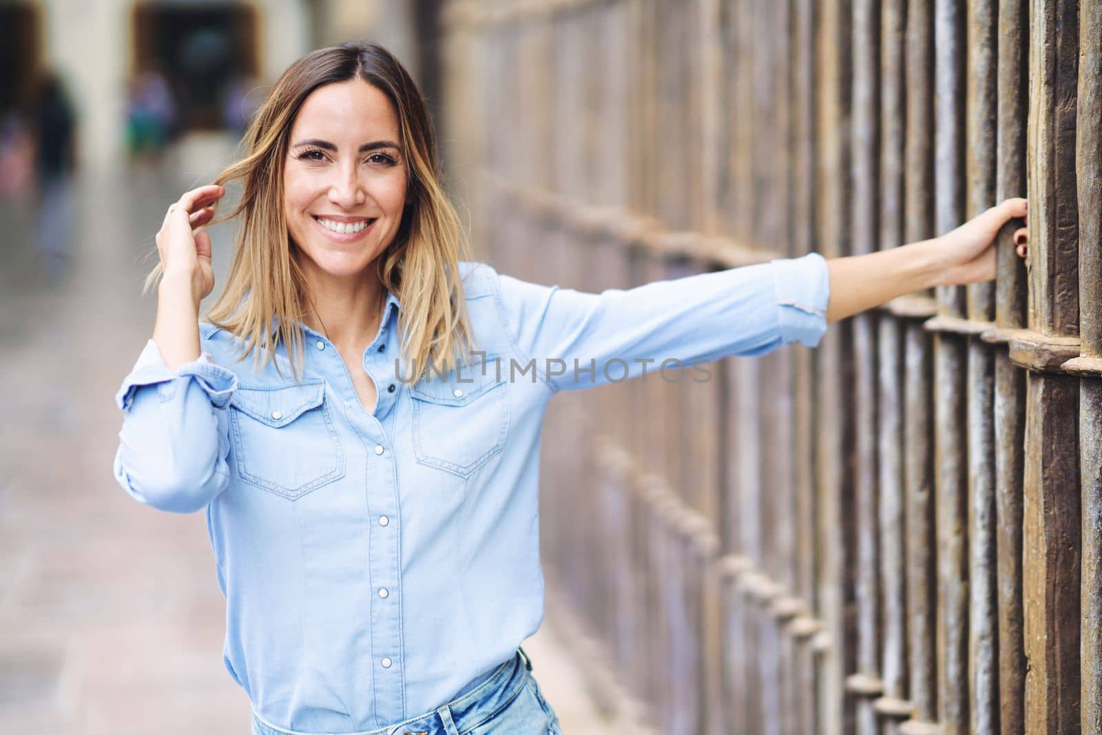 Happy adult female in stylish blue shirt looking at camera with smile and touching hair while grasping metal fence on blurred background of city street