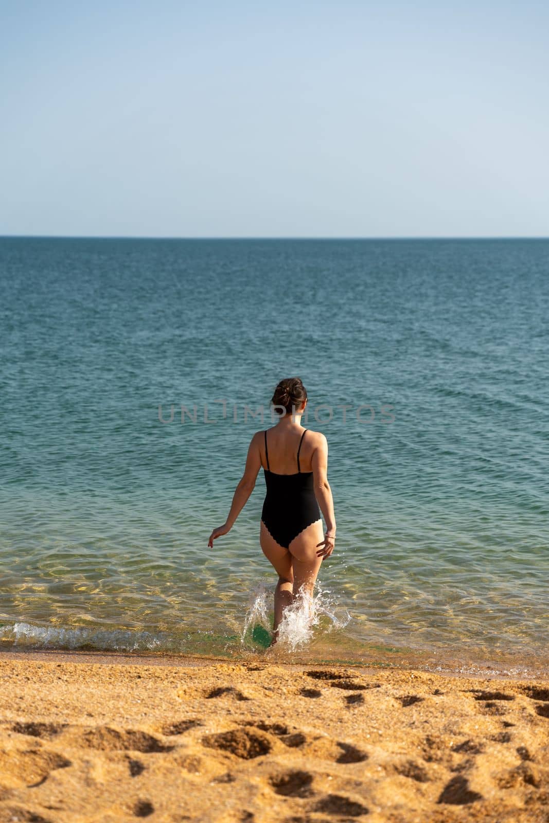 Woman sea swimsuit sand. A girl in full growth stands with her back and enters the sea in a black swimsuit. Alone on the beach on a sunny day. by Matiunina