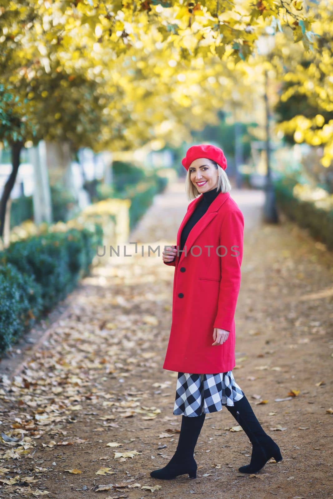 Full body of cheerful female in red clothes looking at camera while standing on pathway in park on autumn day