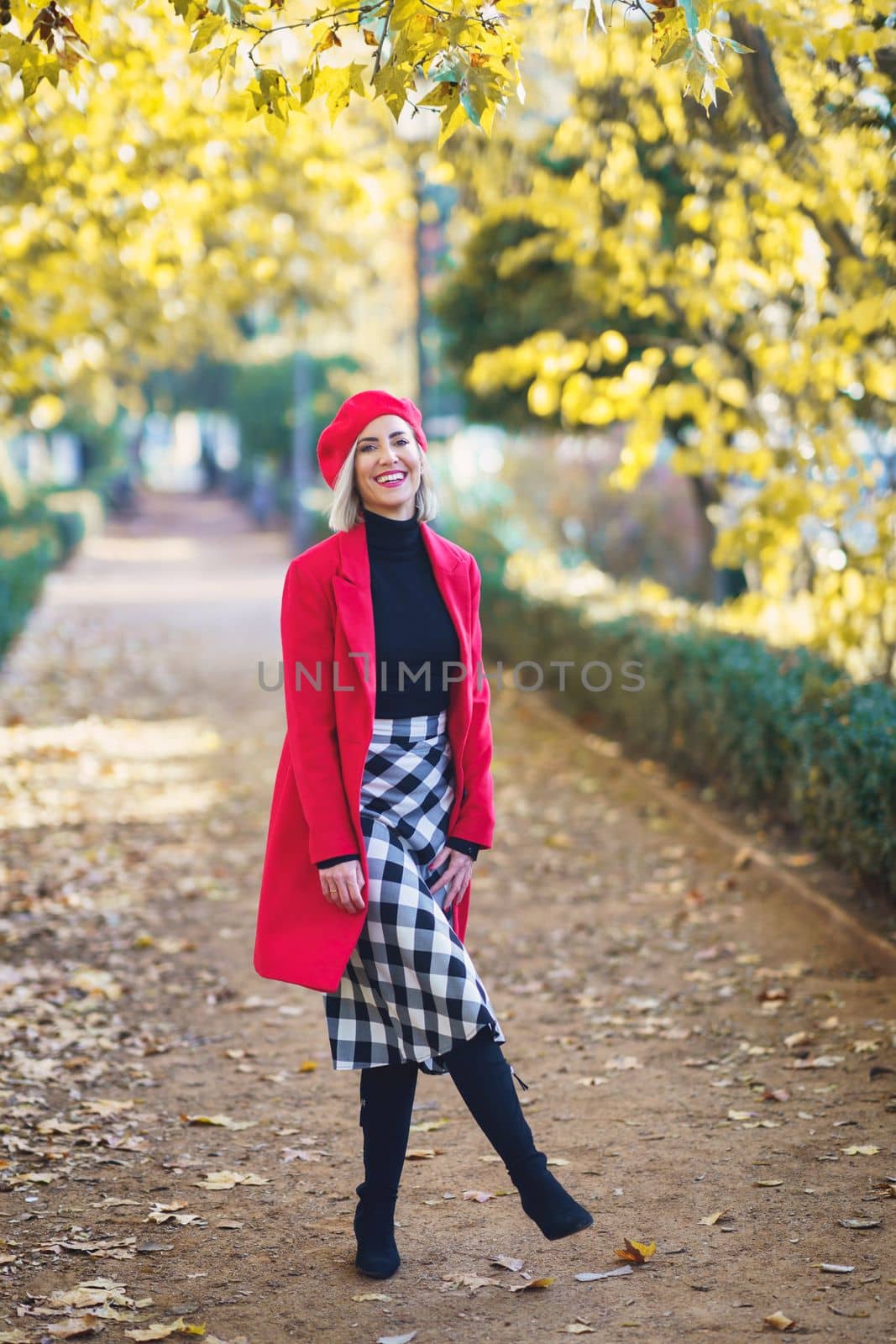 Full body of positive female in stylish outfit looking at camera while standing on pathway in autumn park with trees