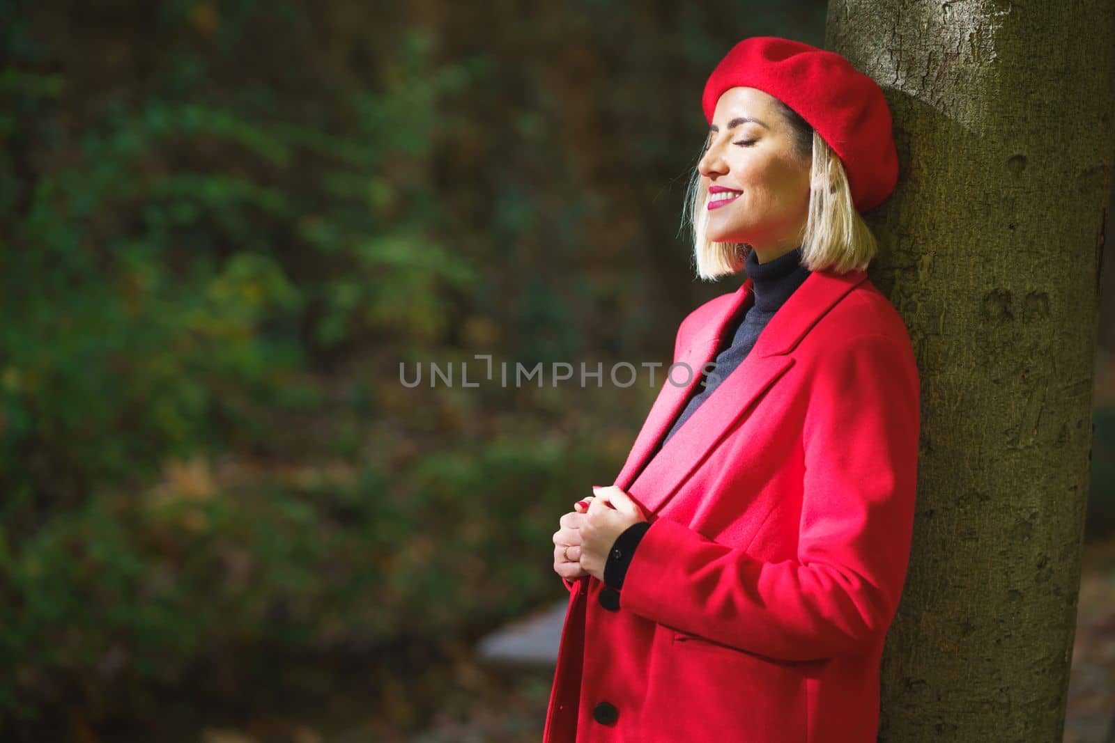 Side view of stylish female wearing red beret and jacket leaning on tree trunk and closing eyes from sunlight in park