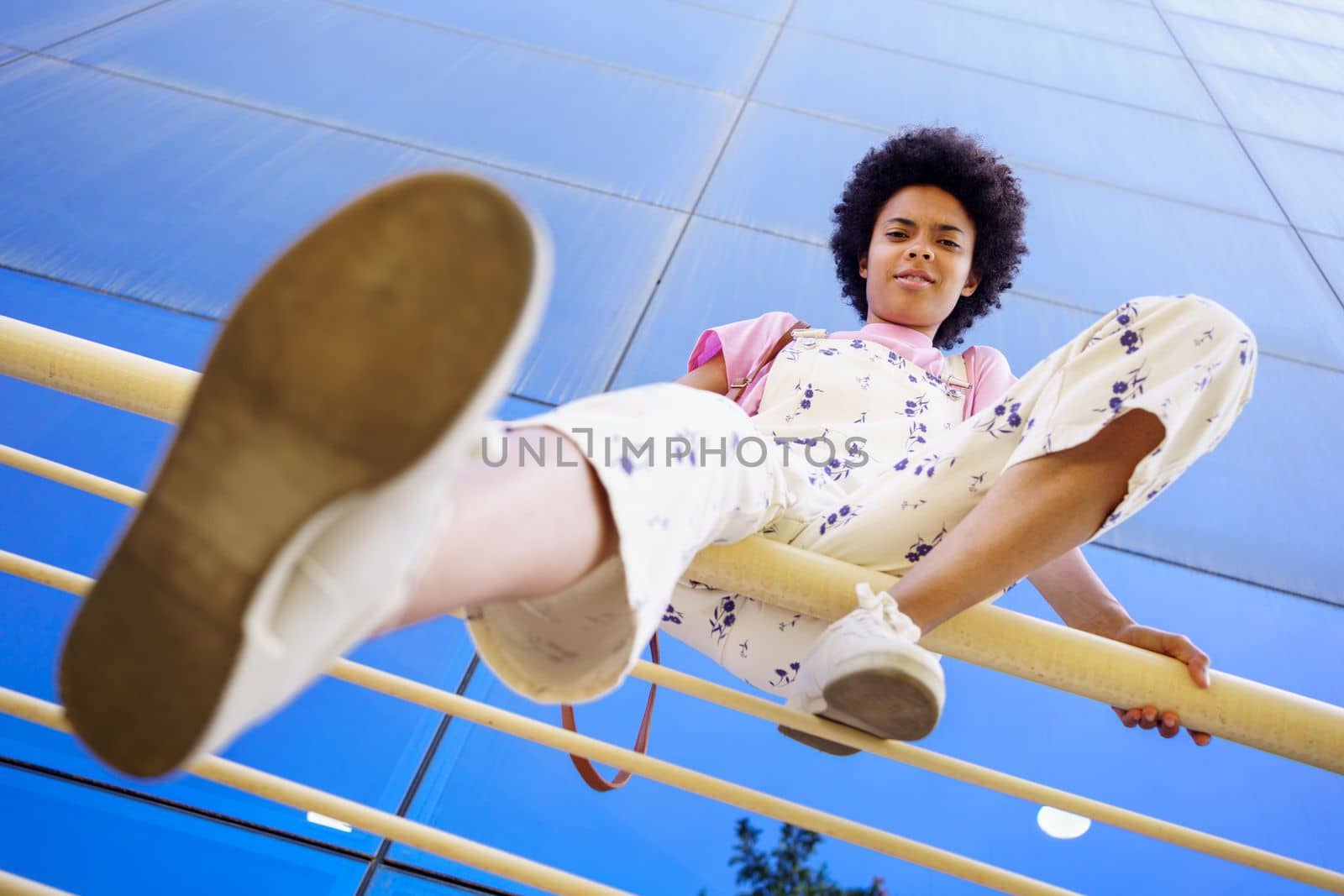 From below full body of young African American female in casual outfit looking at camera with serious face while sitting on railing