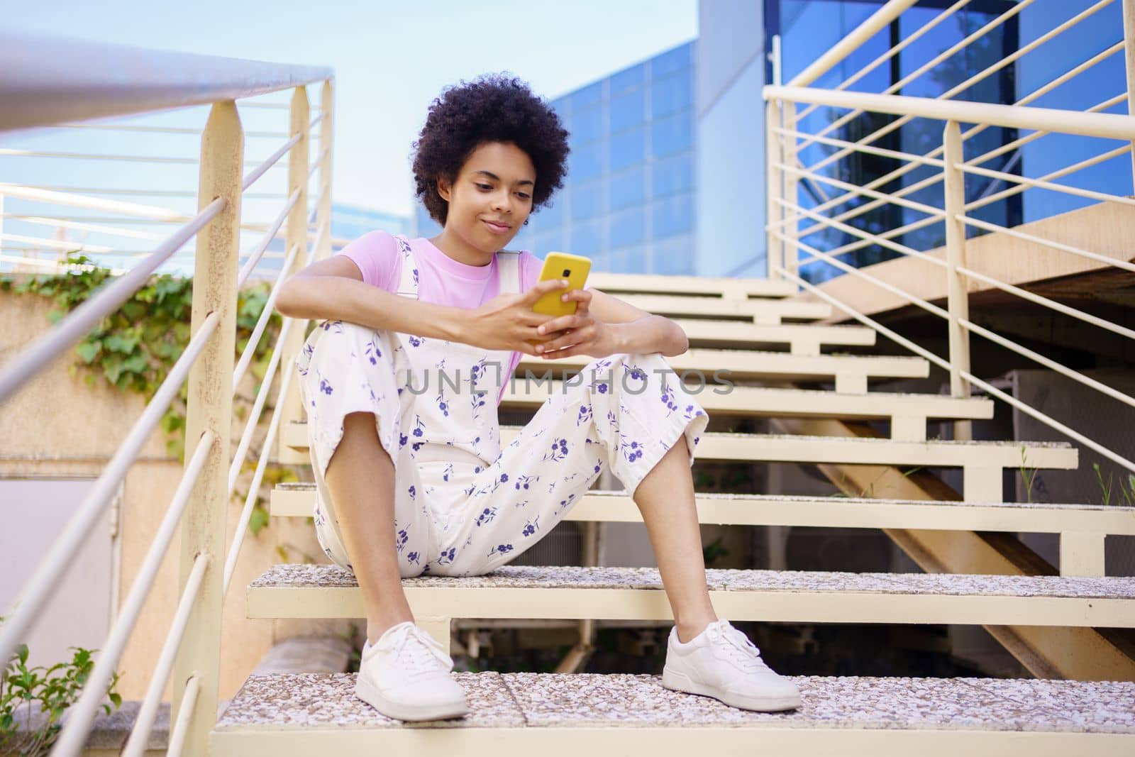 Full body of content African American female in casual wear texting message on cellphone while sitting on stairway in city