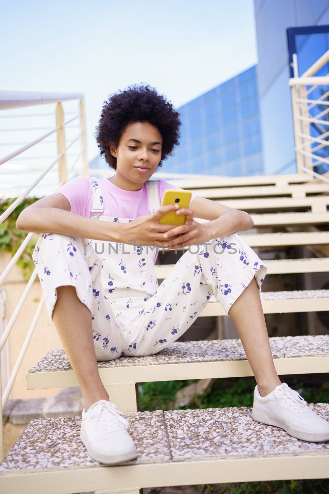 Black woman texting with her smartphone, sitting on steps of a modern building. by javiindy