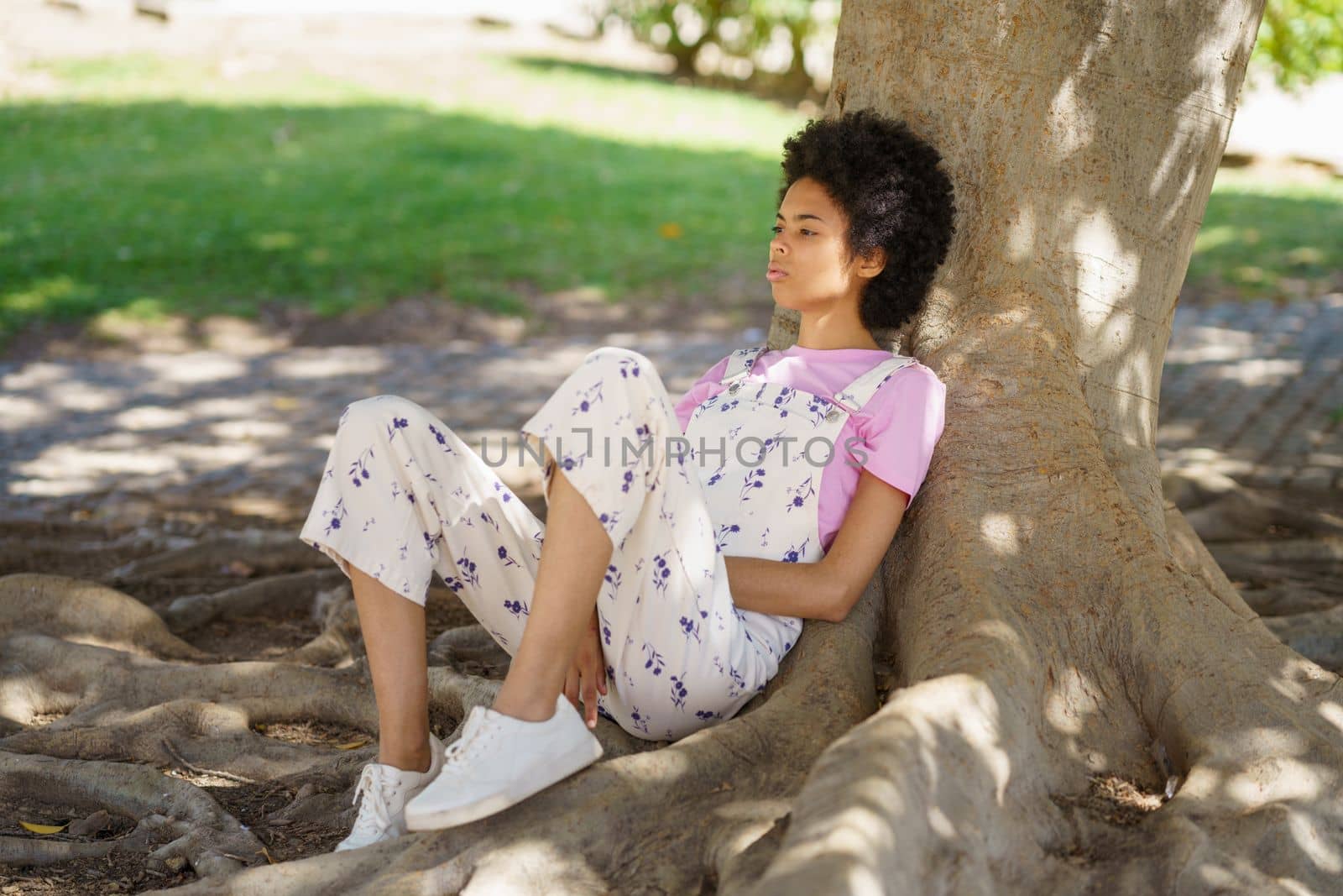 Full body side view of young wistful African American female in casual clothes sitting near thick tree trunk in park