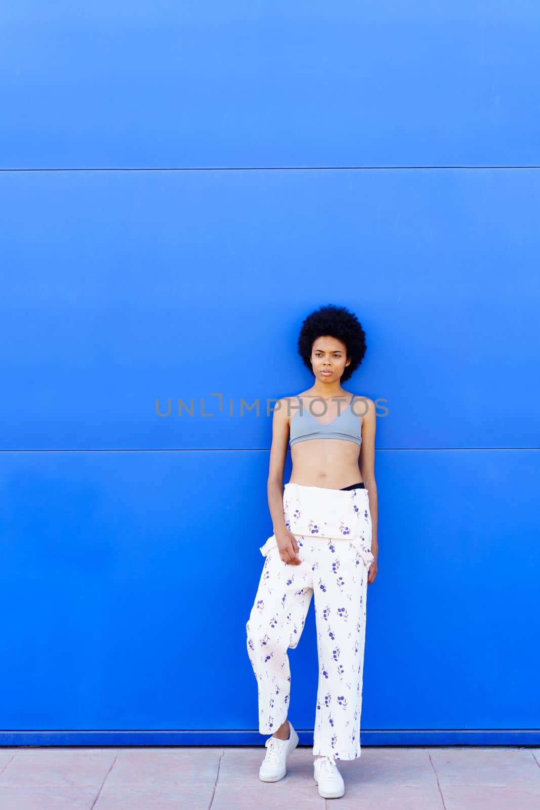 Full body of fit African American female in bra and overall looking away while standing on blue background in city
