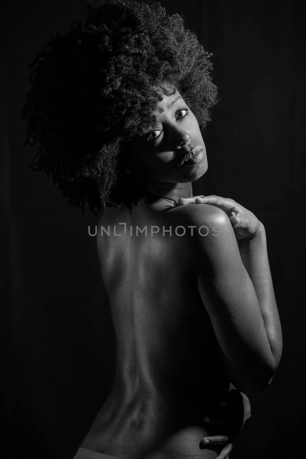 Sensual nude African American female with curly hair looking away over shoulder while standing against black background. Black and white photograph.
