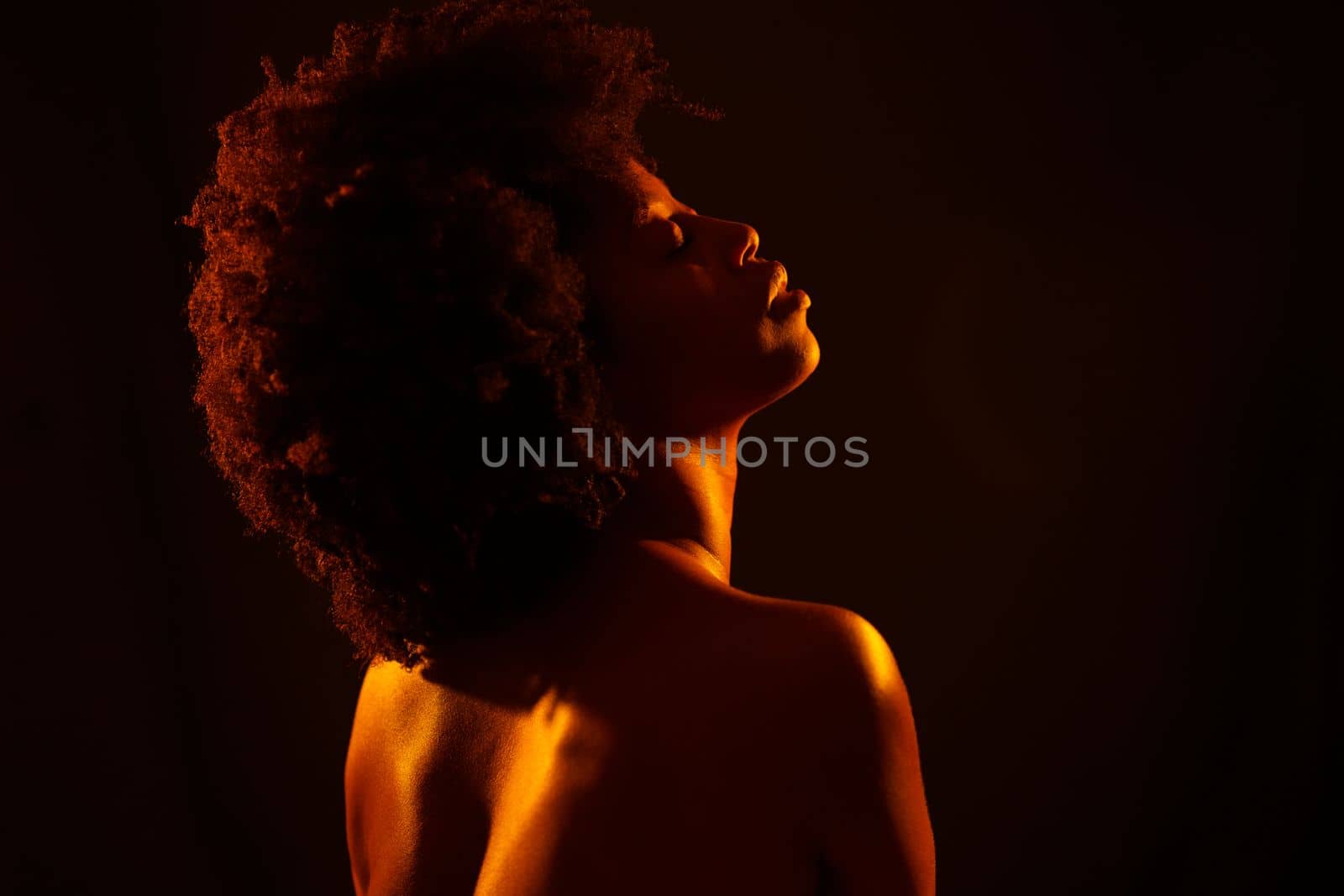 Naked black woman model with Afro hairstyle closing eyes and enjoying bright neon orange light against black background