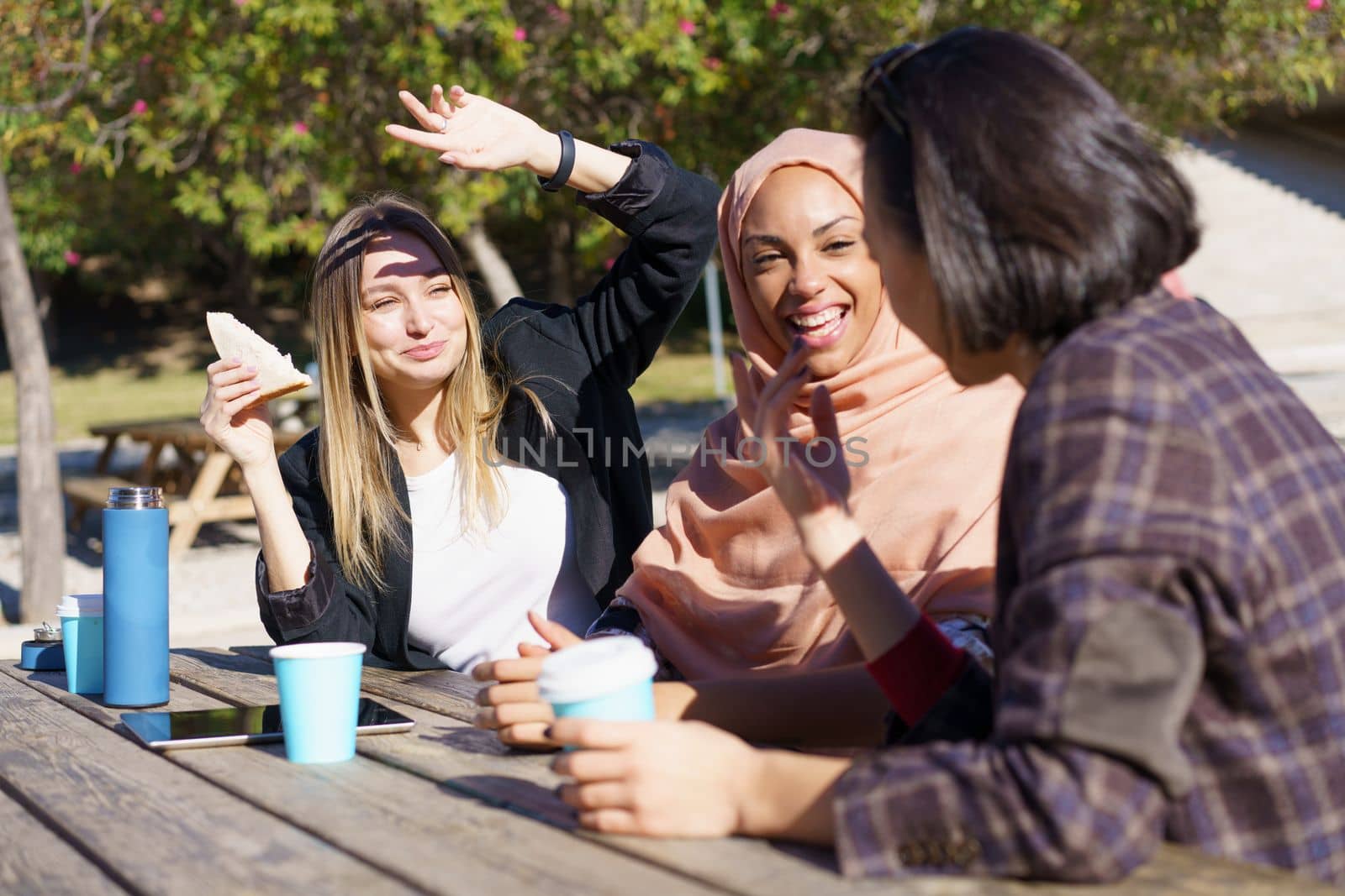 Delighted multiethnic female friends drinking takeaway coffee and smiling in park by javiindy