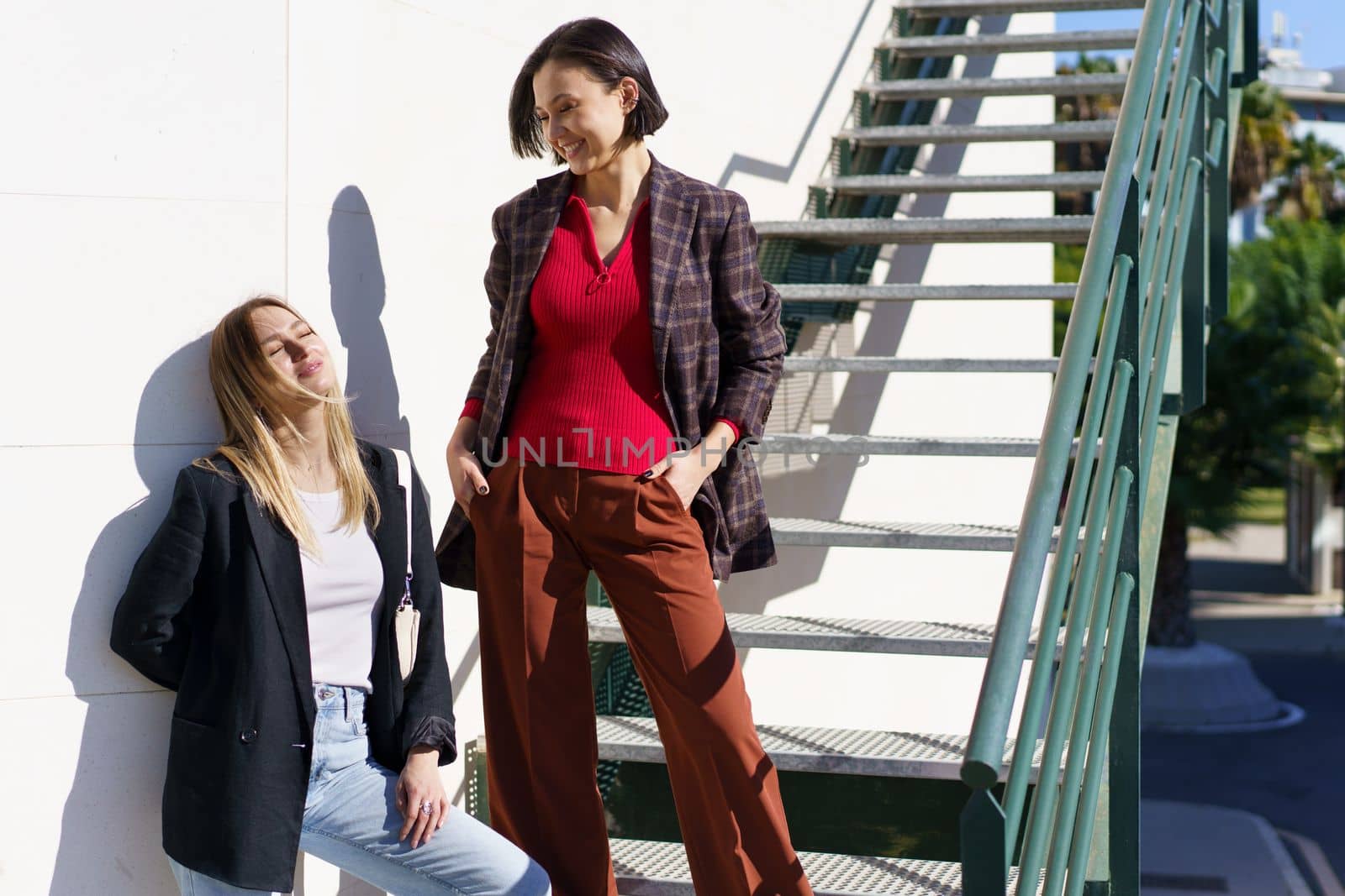 Content young women best friends in stylish outfits enjoying sunny day standing on staircase in city park