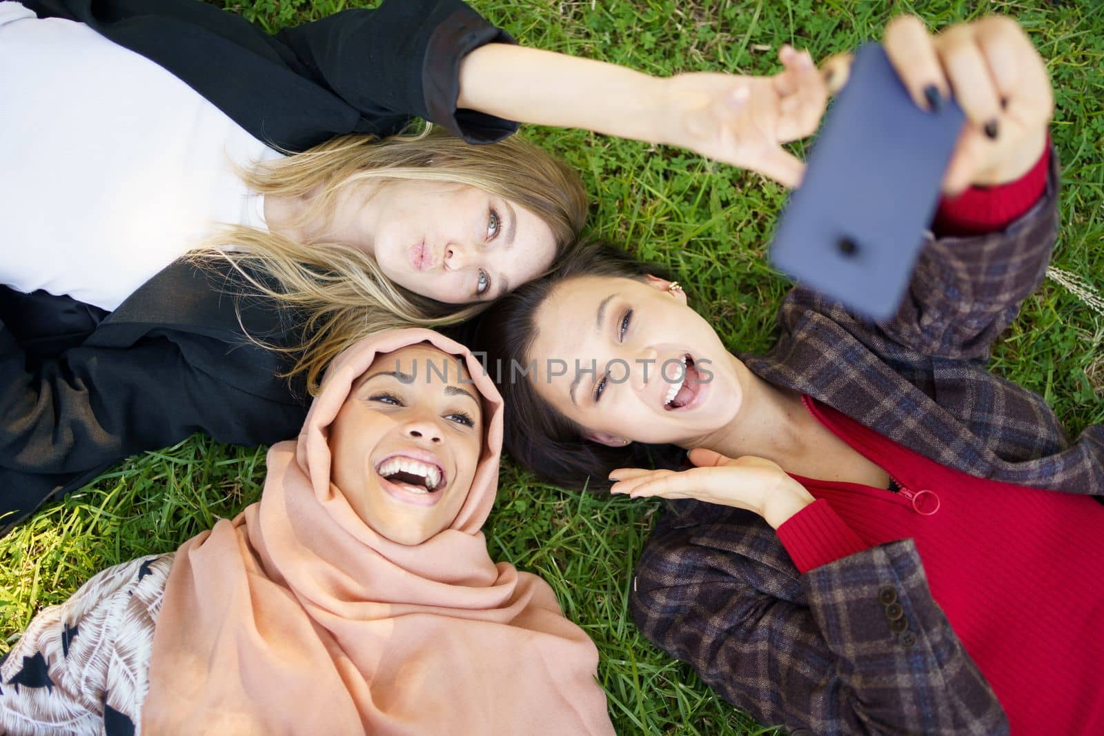 Top view of laughing Muslim female lying near girlfriends pouting lips and taking self portrait on cellphone on grassy ground