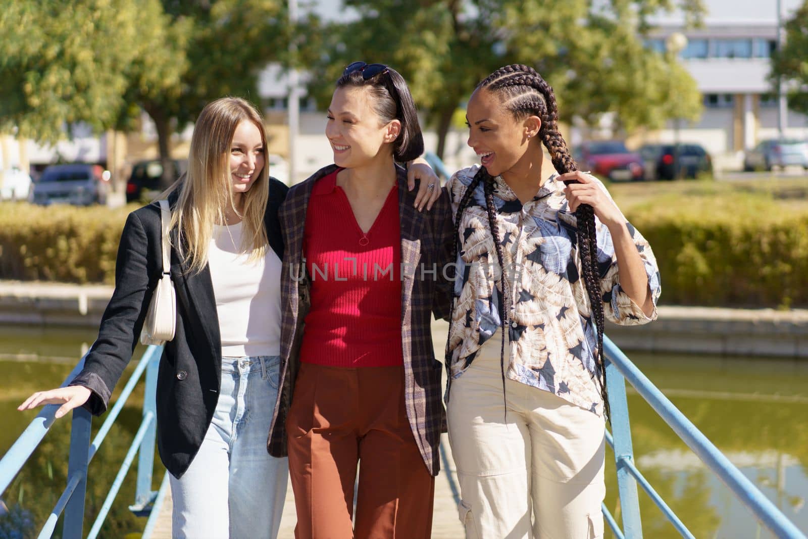 Group of content diverse female friends strolling on footbridge crossing river on sunny summer day in city with green trees