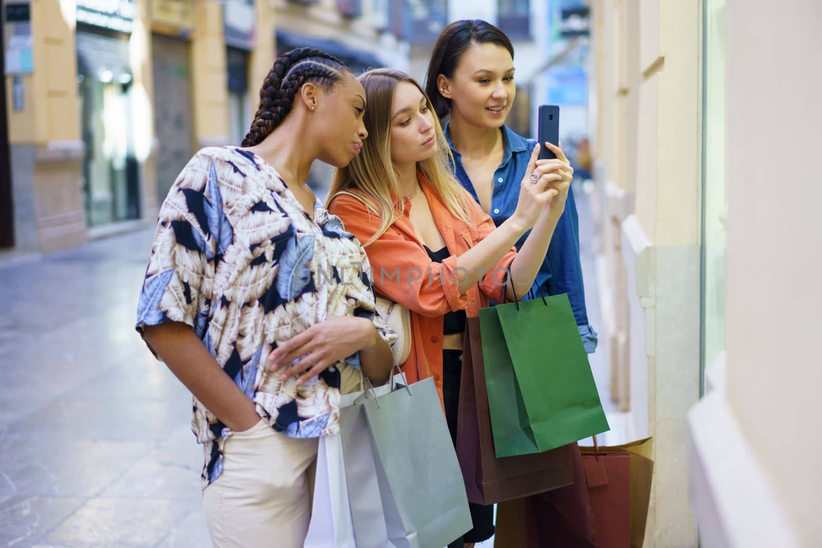 Group of diverse female friends with shopping bags taking picture of showcase and choosing goods in store while standing on street
