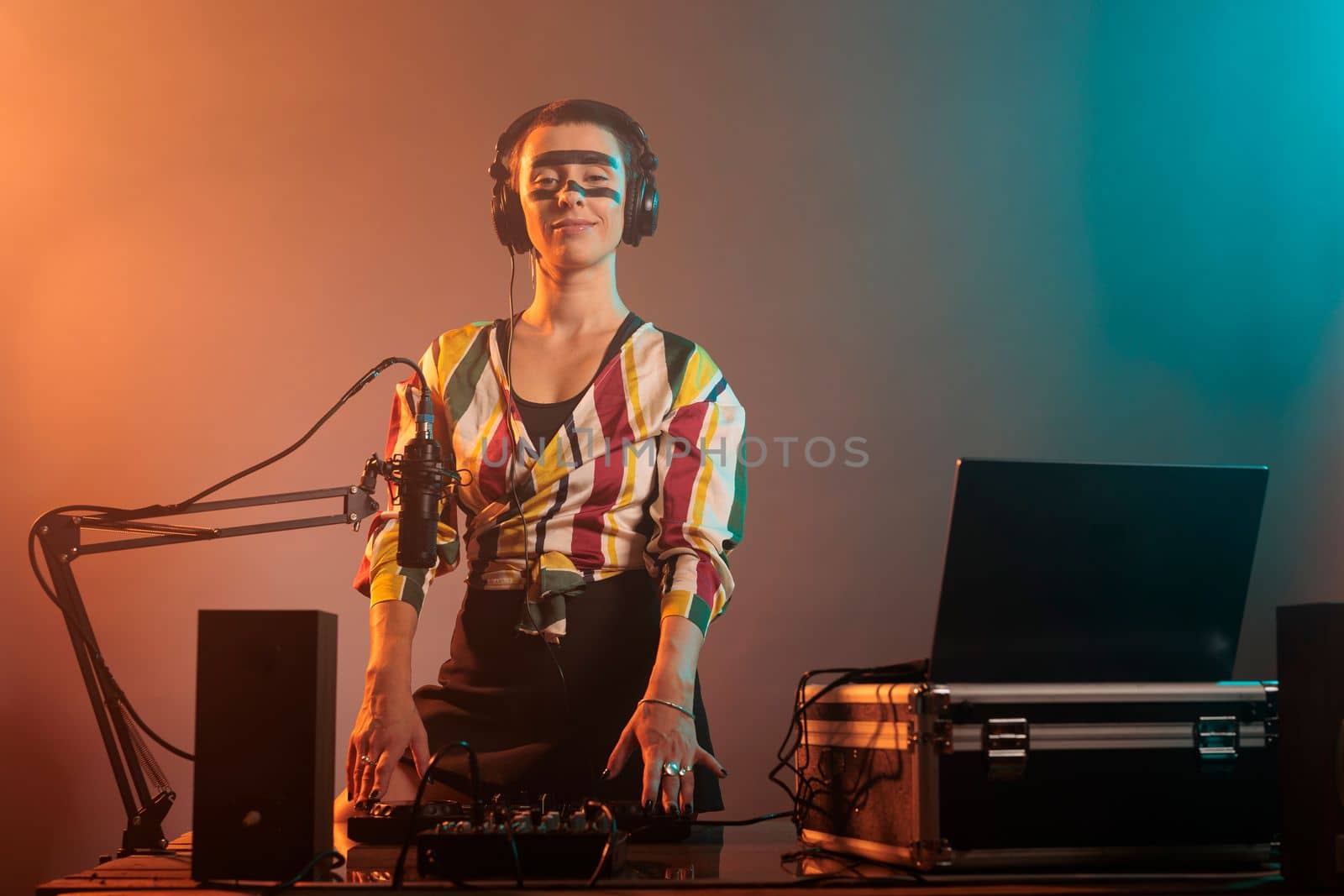 Happy female performer working as dj with turntables, mixing techno music with bass and audio equipment. DJ woman playing songs at mixer, standing over colorful background with smoke.