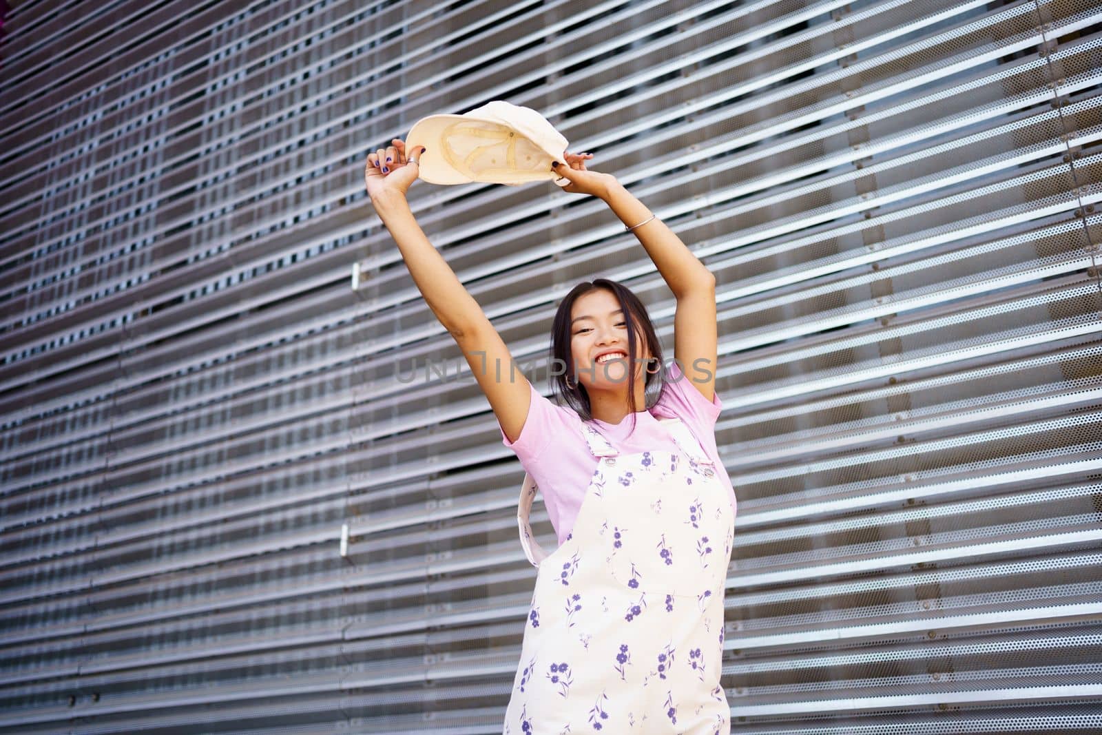 Low angle of optimistic young Asian woman in stylish clothes, raising arms with cap and looking at camera with smile against metal wall in daytime on city street