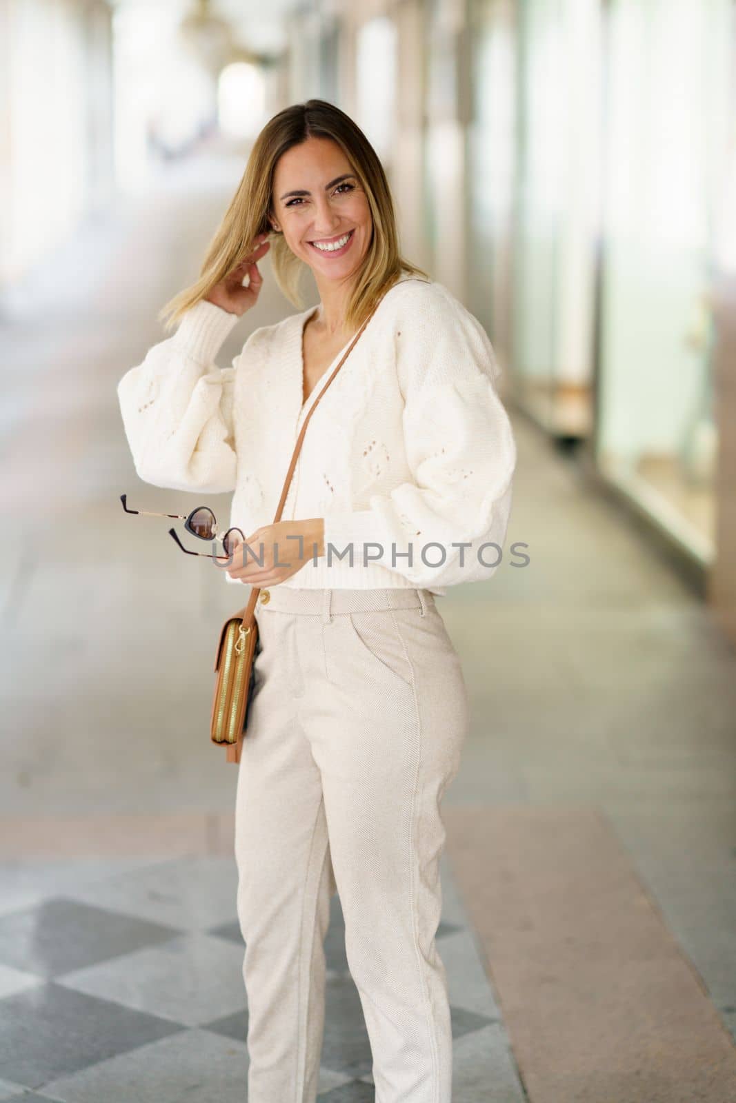 Positive young blond haired female in smart casual outfit smiling and looking at camera while fixing hair