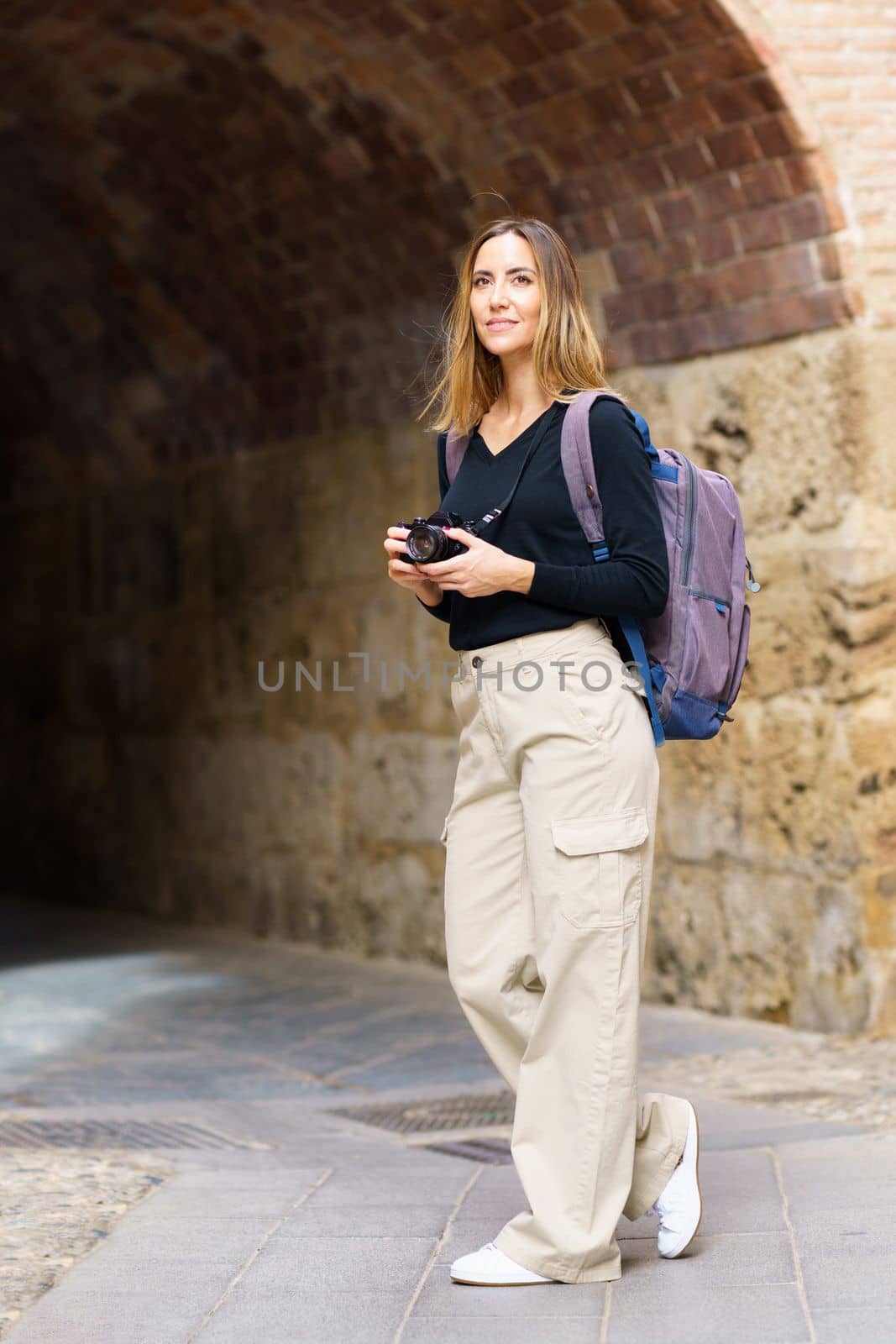 Side view of positive young female tourist with blond hair in casual wear and backpack, using photo camera and smiling while standing near ages stone archway during sightseeing trip