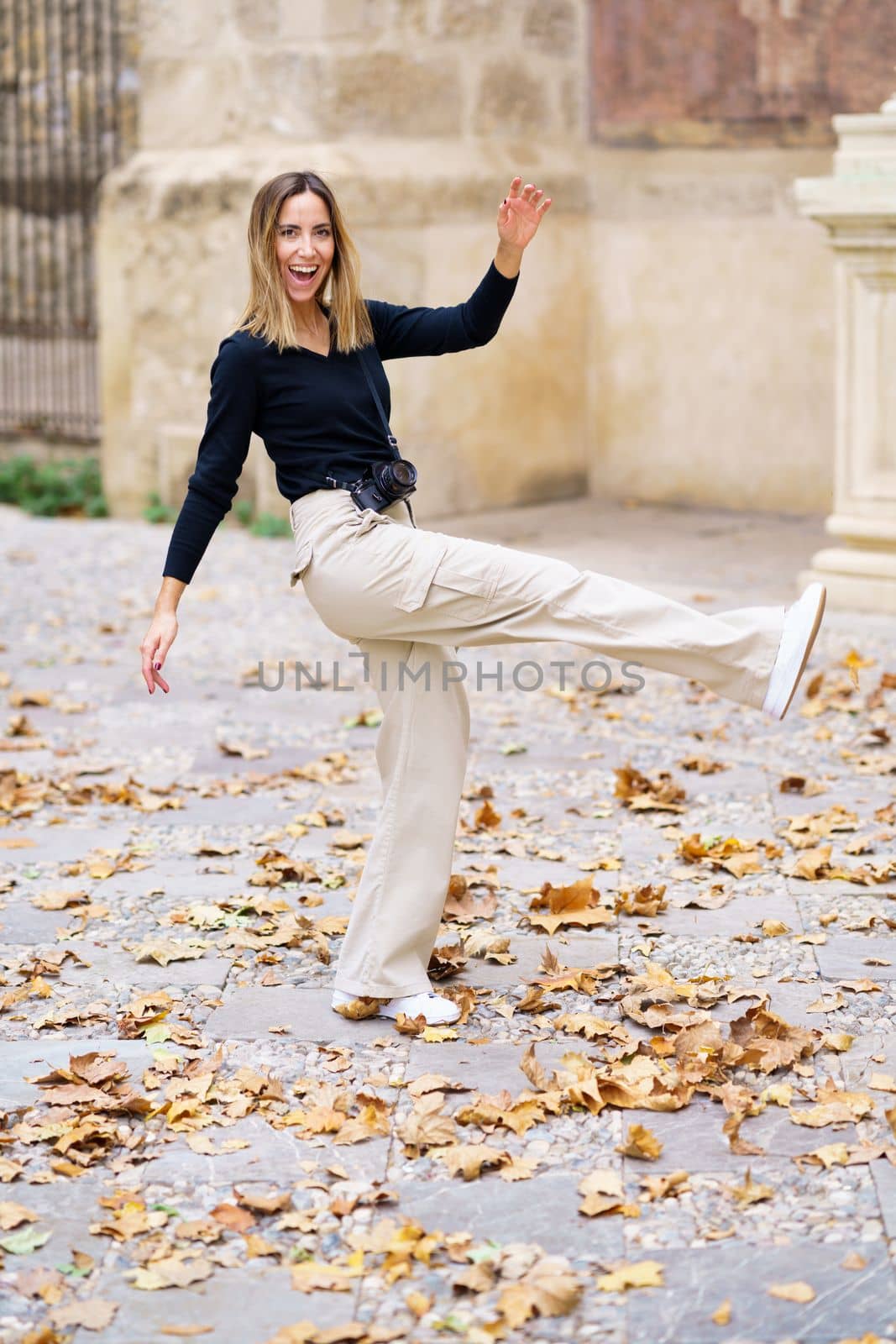 Side view of delighted young female tourist with long blond hair in casual clothes, smiling brightly and looking at camera while kicking golden fallen leaves during autumn holidays