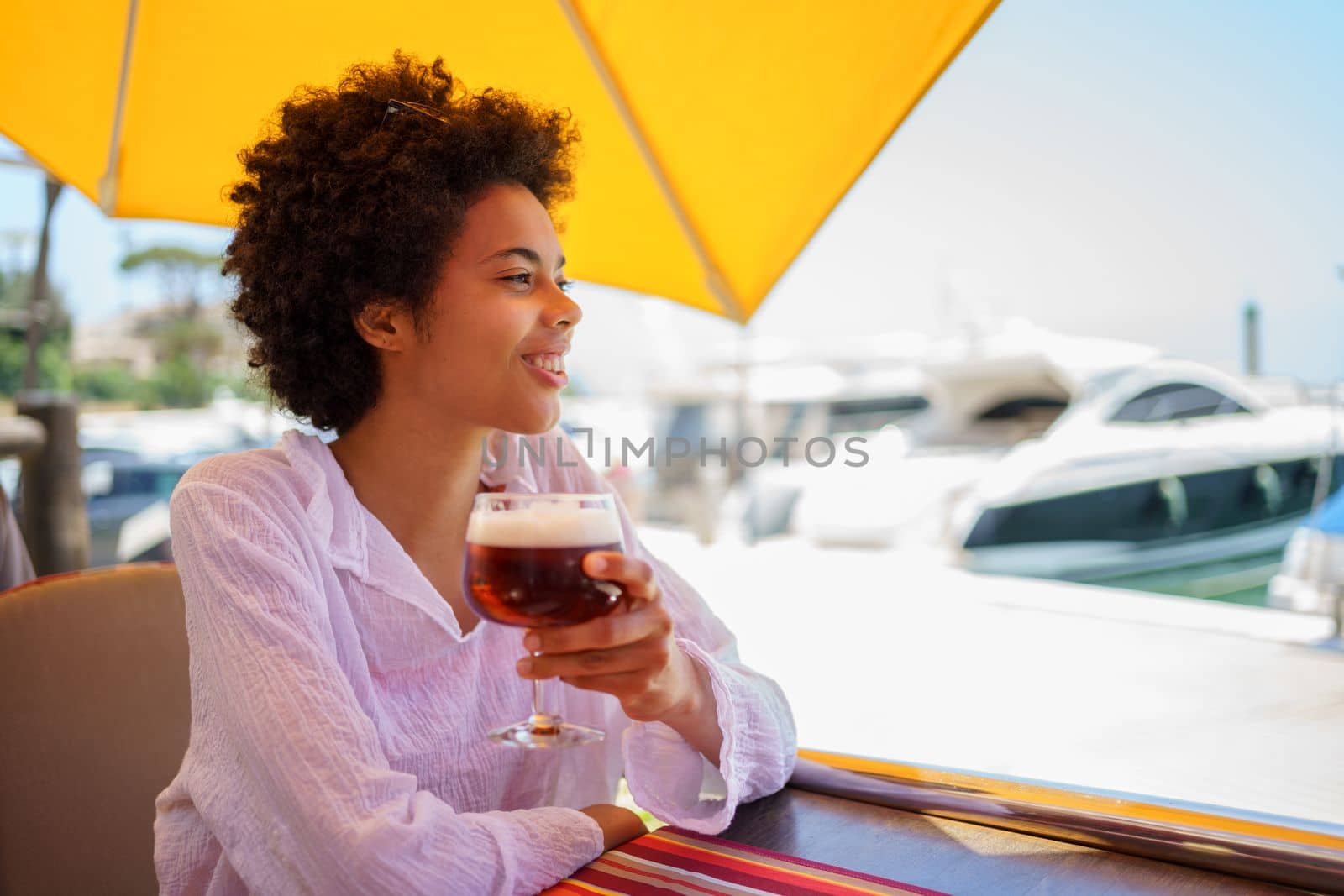 Cheerful young African American female in white blouse with curly hair smiling and looking away, while enjoying beer in street restaurant on sunny summer day on embankment