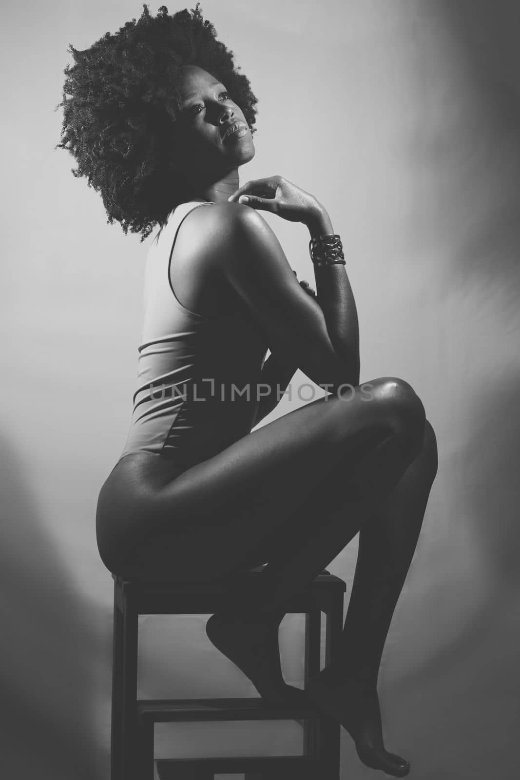 Side view of slim African American female model in bodysuit, with curly hair touching shoulder and looking away while sitting on stool. Black and white photograph.